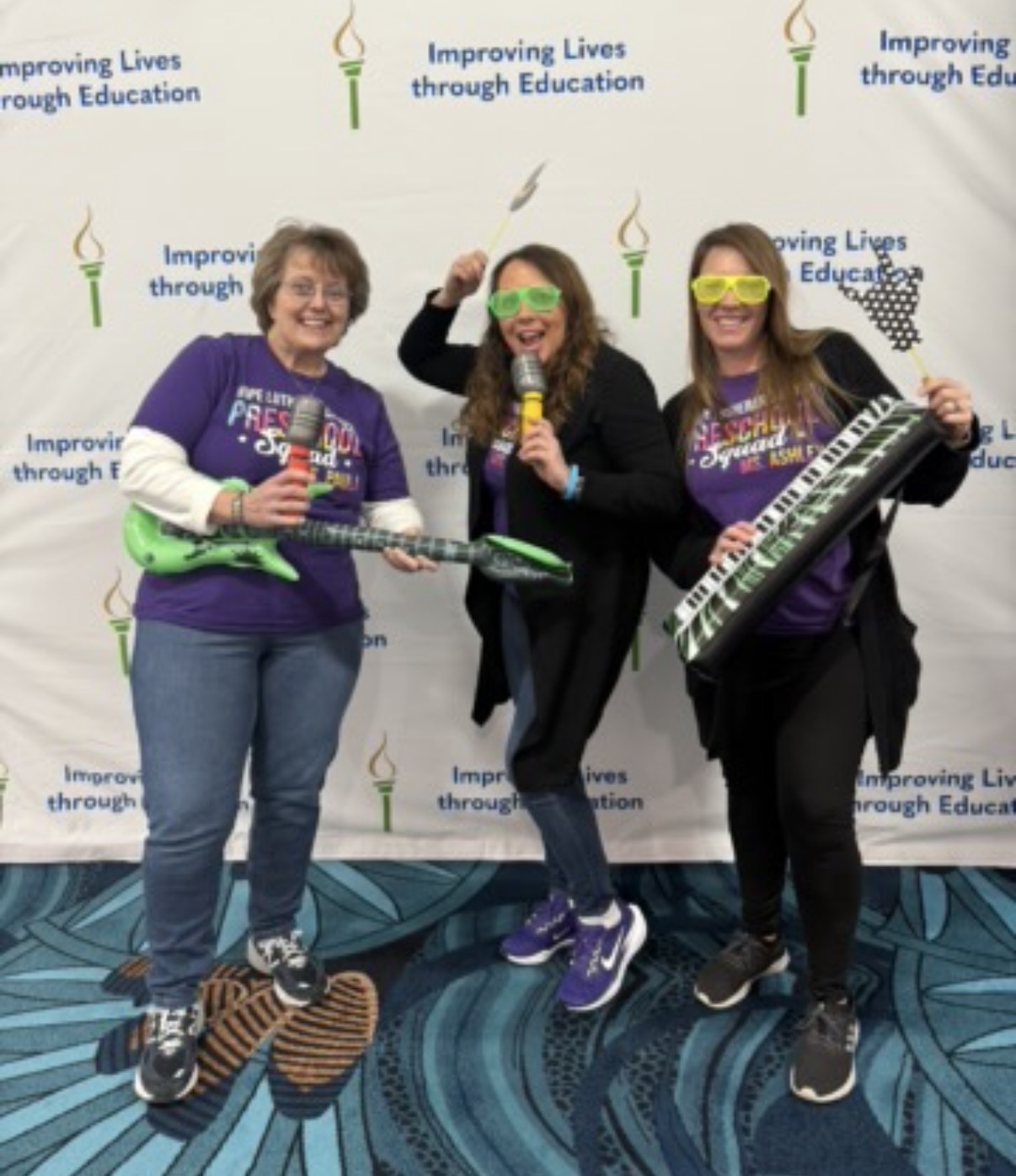 Three women smiling, holding musical instruments, standing in front of a backdrop with educational logos, on a patterned blue carpet.