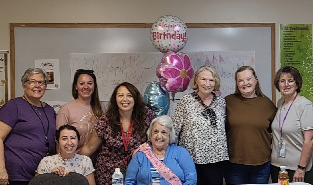 Group of eight women celebrating a birthday with balloons and a birthday sash, standing in front of a whiteboard.