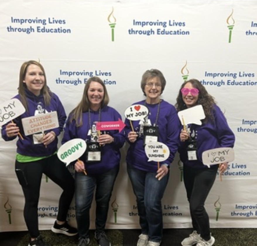 Four women stand in front of a backdrop with educational slogans, holding signs and smiling.