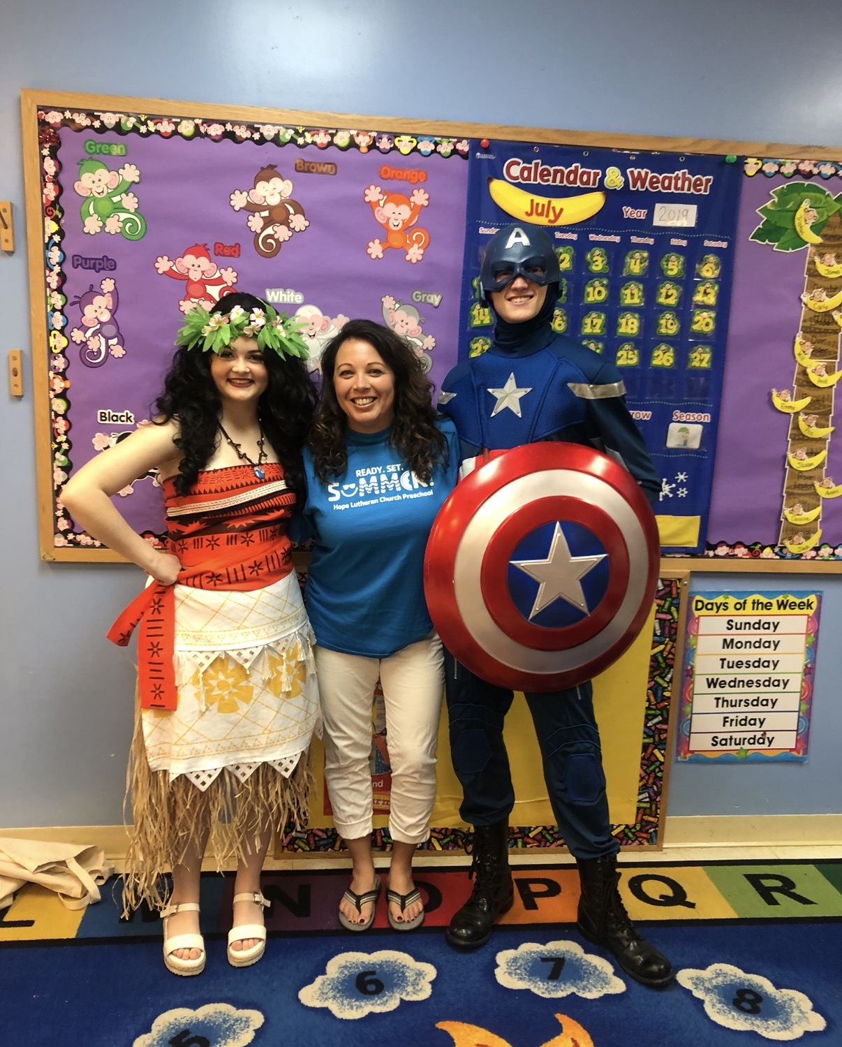 Three people in costumes stand in front of a classroom bulletin board, smiling. One is dressed as a hula dancer, another as a woman in casual clothes, and the third as Captain America holding a shield.