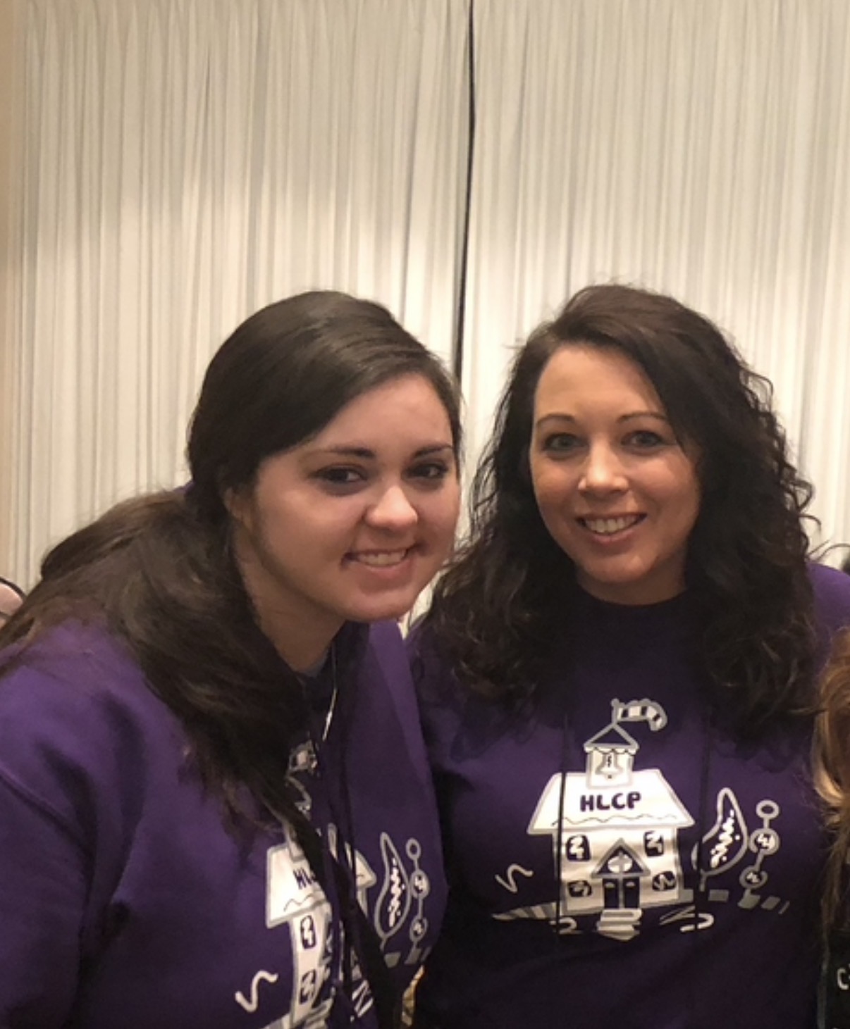 Two women smiling, wearing purple shirts with a boat logo, standing in front of a curtain backdrop.