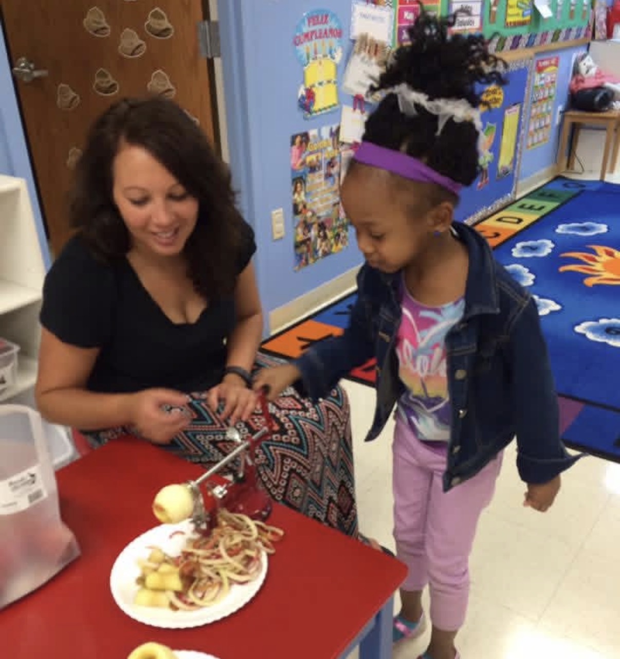 Two girls, one adult and one child, smiling and interacting with a plate of food on a red table in a colorful classroom.
