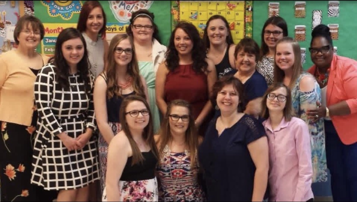 Group of fifteen women and girls standing in a classroom with colorful posters and decorations behind them.