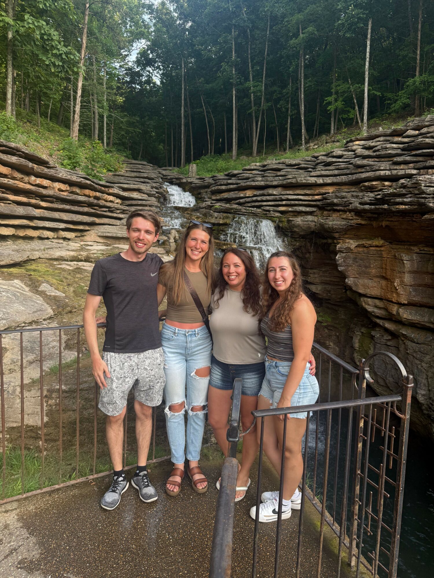 Three women and one man standing outdoors on a rocky area with trees in the background, smiling at the camera.