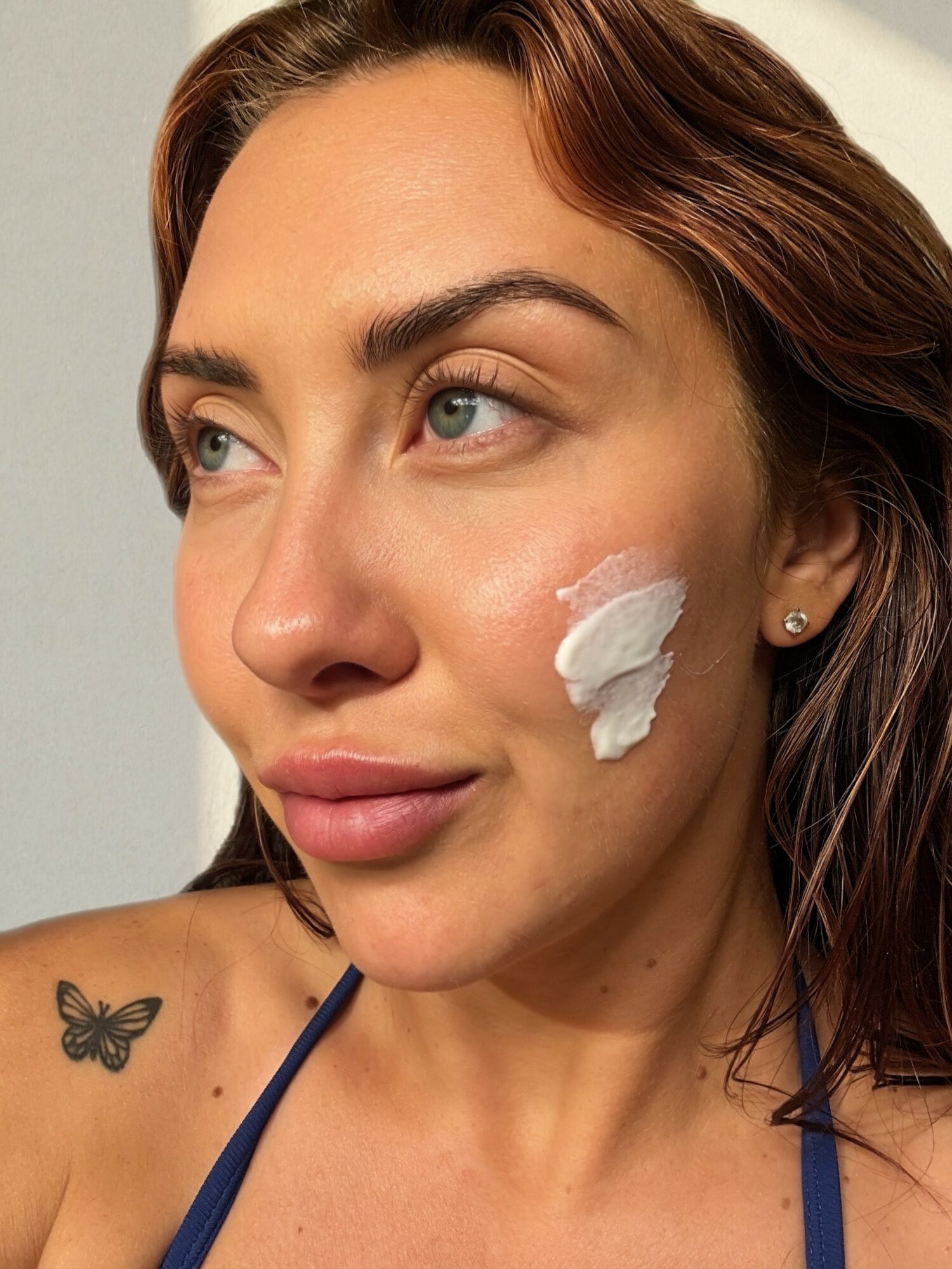 Woman with light skin and brown hair, applying cream to her cheek, butterfly tattoo on shoulder, wearing a blue strap top.