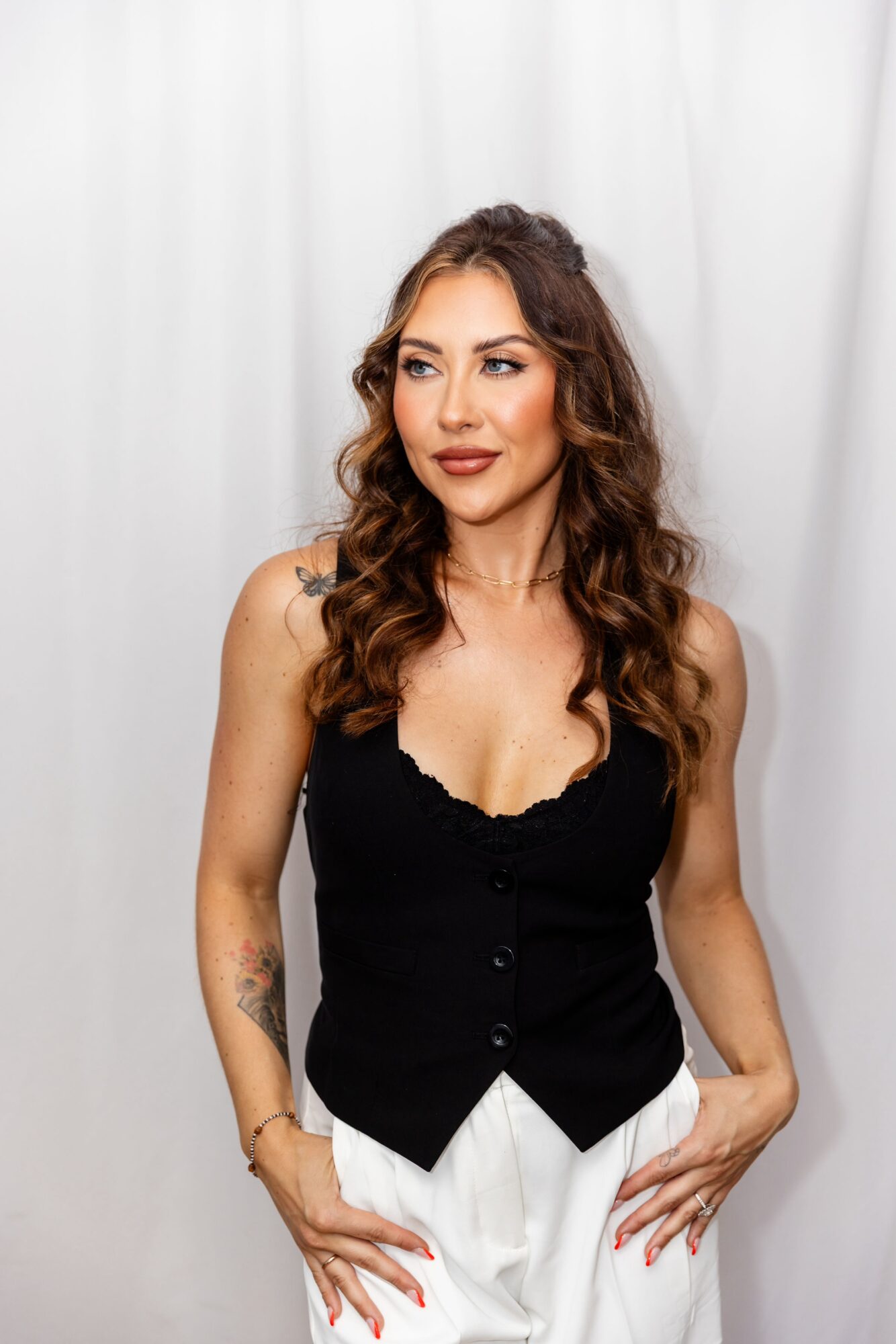 Woman with wavy brown hair wearing a black sleeveless top and white pants, standing against a white background.