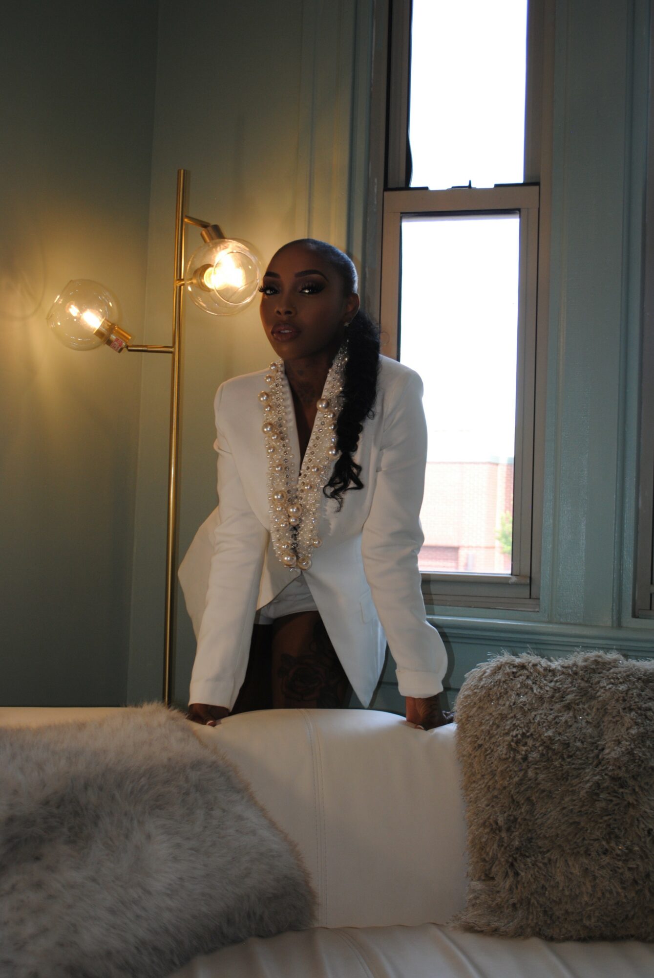 Woman in white blazer standing near window with green walls and a wall-mounted lamp, looking at camera, with pillows in foreground.