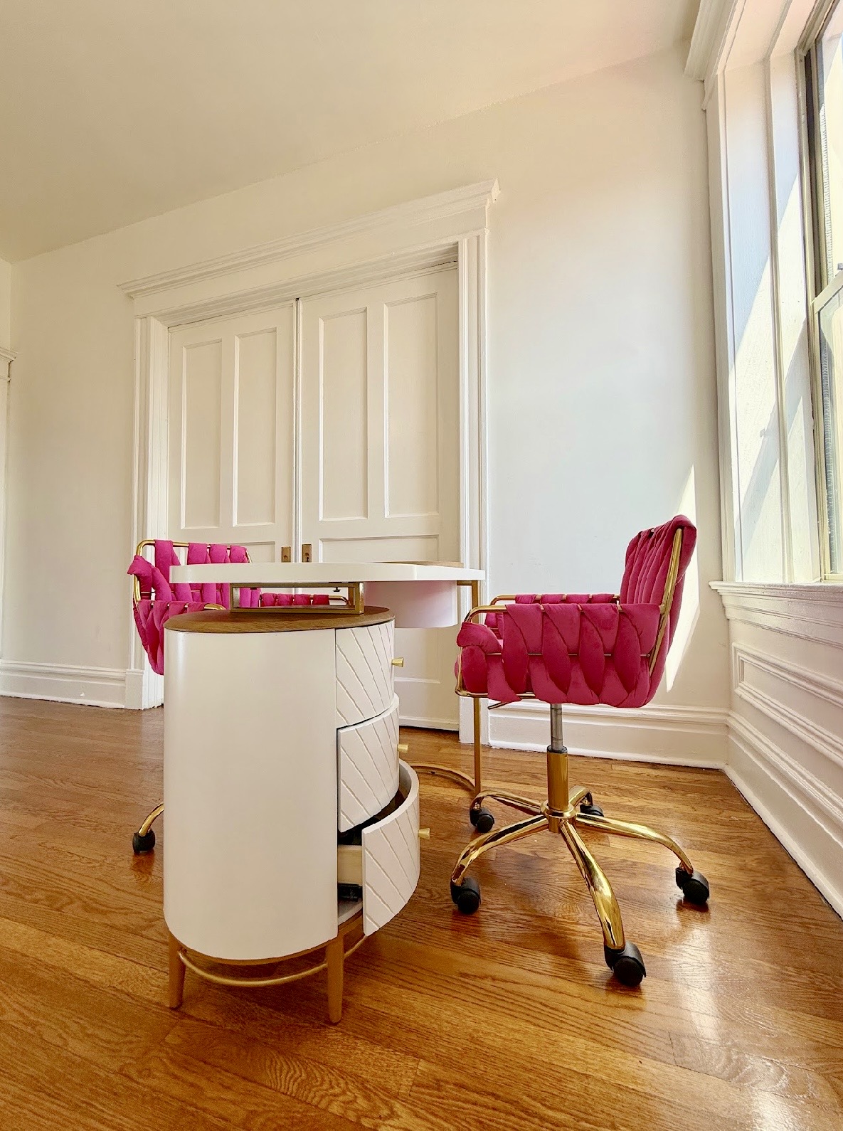 Corner of a room with a white cabinet, pink chair, and a pink office chair near a window, hardwood floor, and white walls.