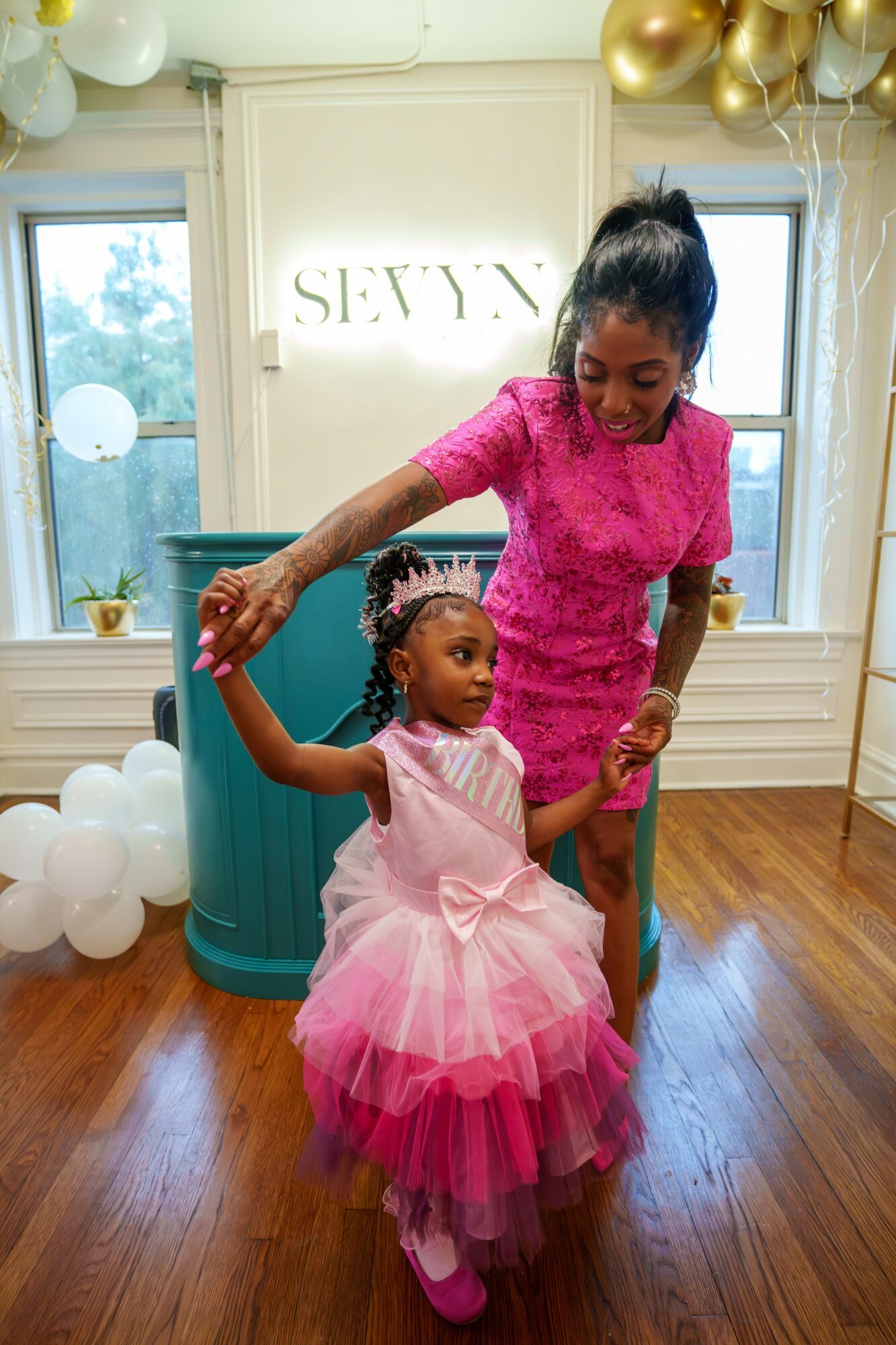 Woman and girl dancing indoors with balloons and decorations, girl in pink dress, woman in pink lace dress, wooden floor, window in background.