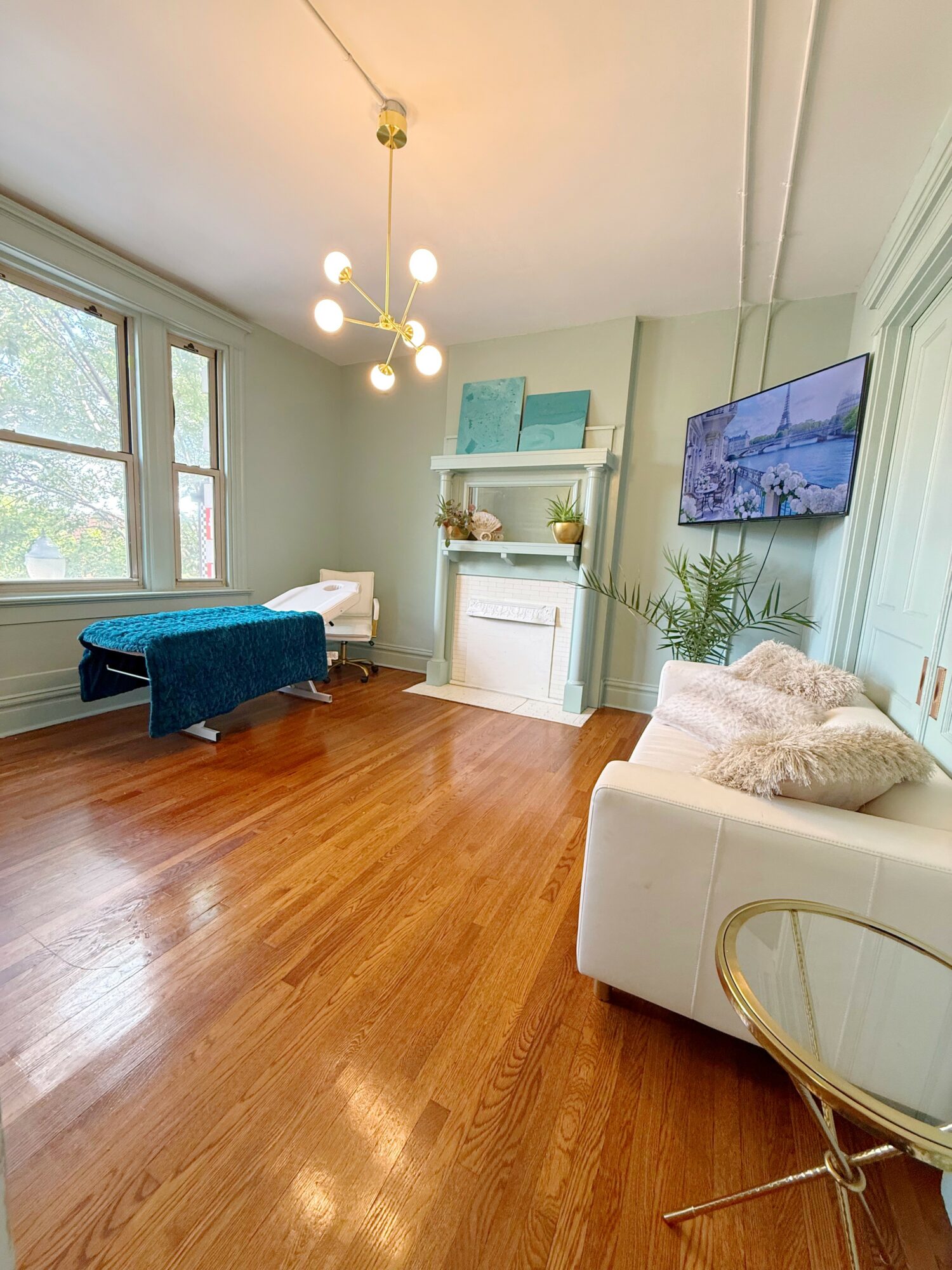 Living room with large window, white sofa, wall-mounted TV, wooden floor, and decorative shelf with artwork.