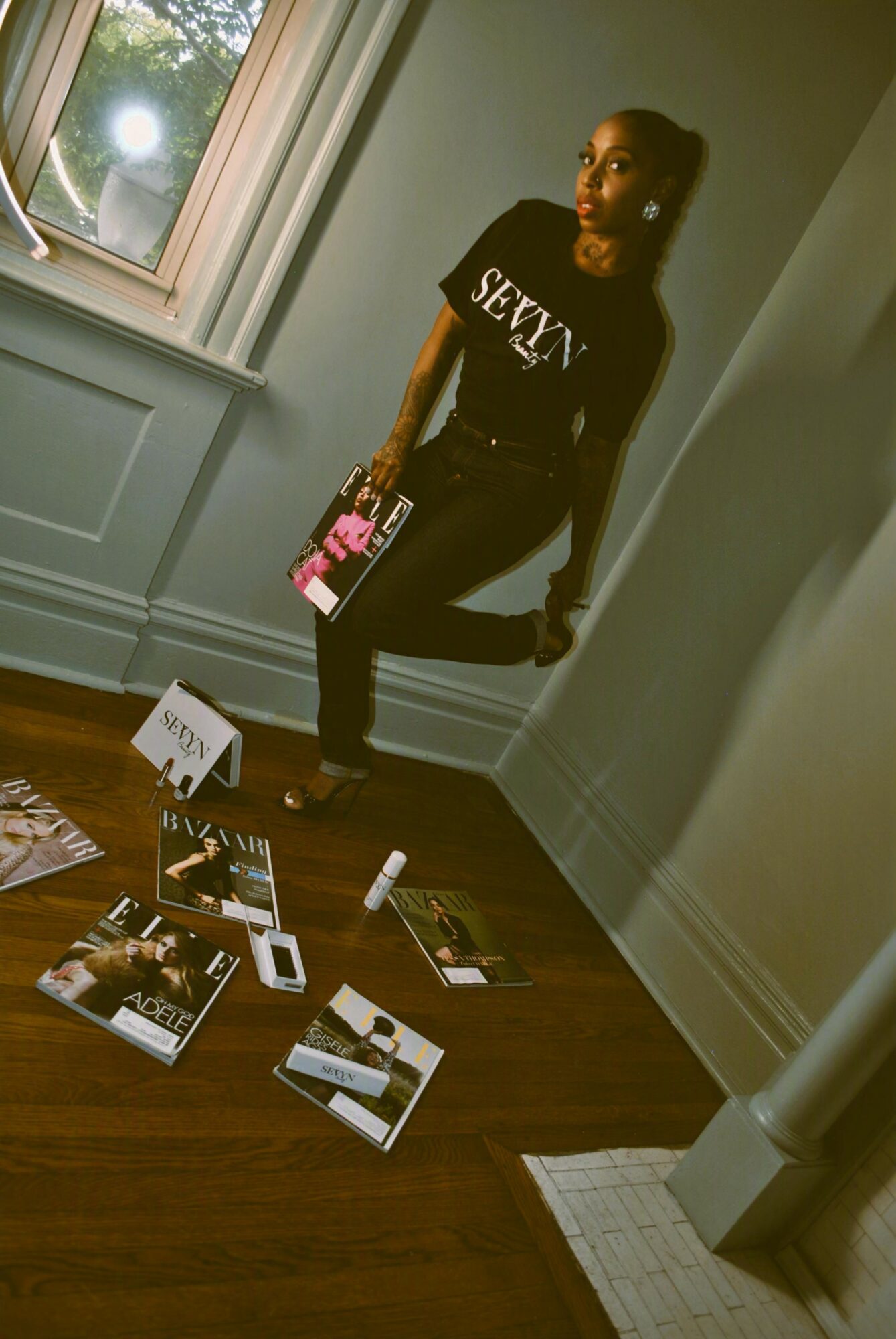 Woman sitting on a windowsill in a room with magazines and makeup on the floor, wearing a black shirt with 'SEXY' text.