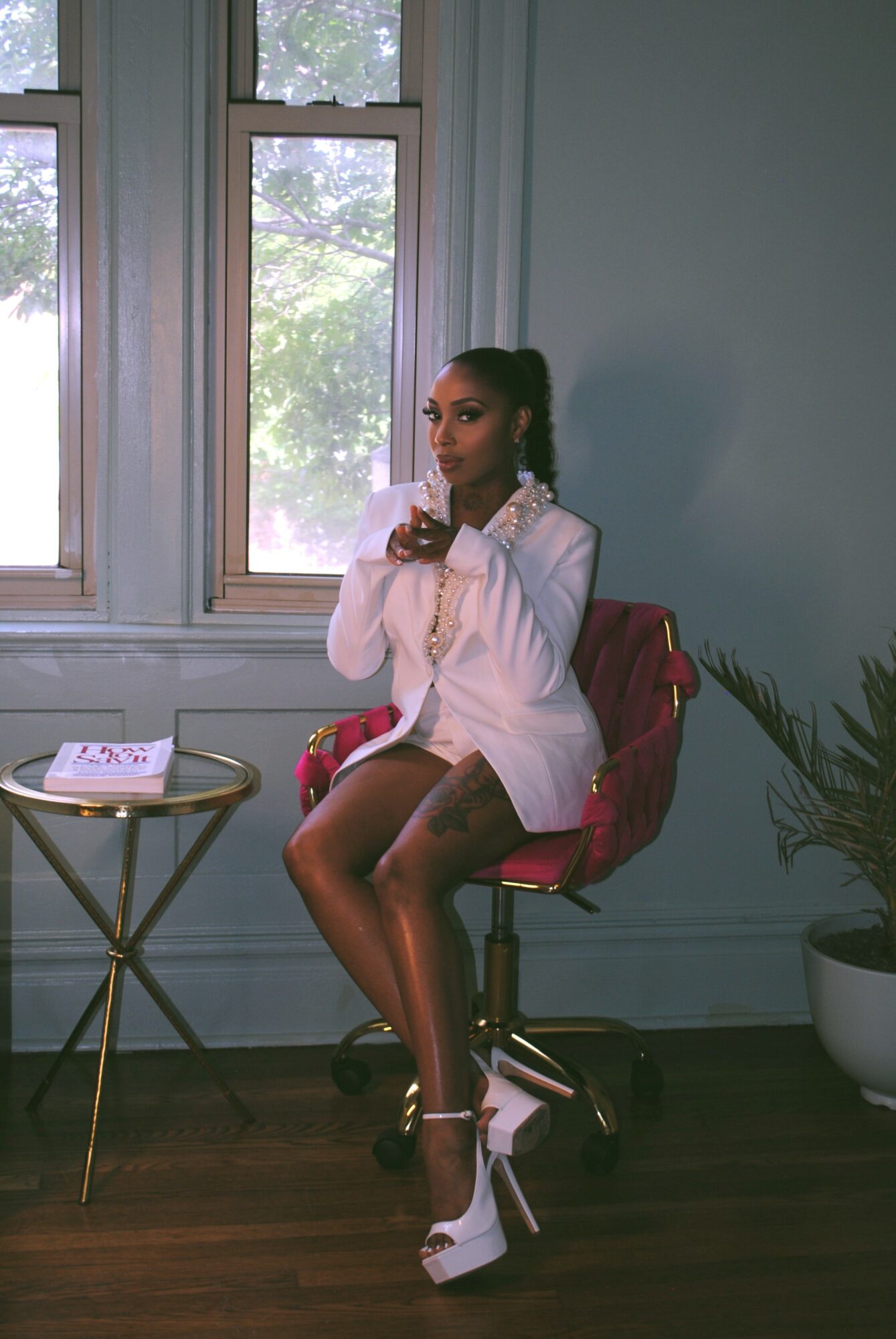 Woman sitting on a red chair near windows, holding a cup, with a small table and potted plant nearby.