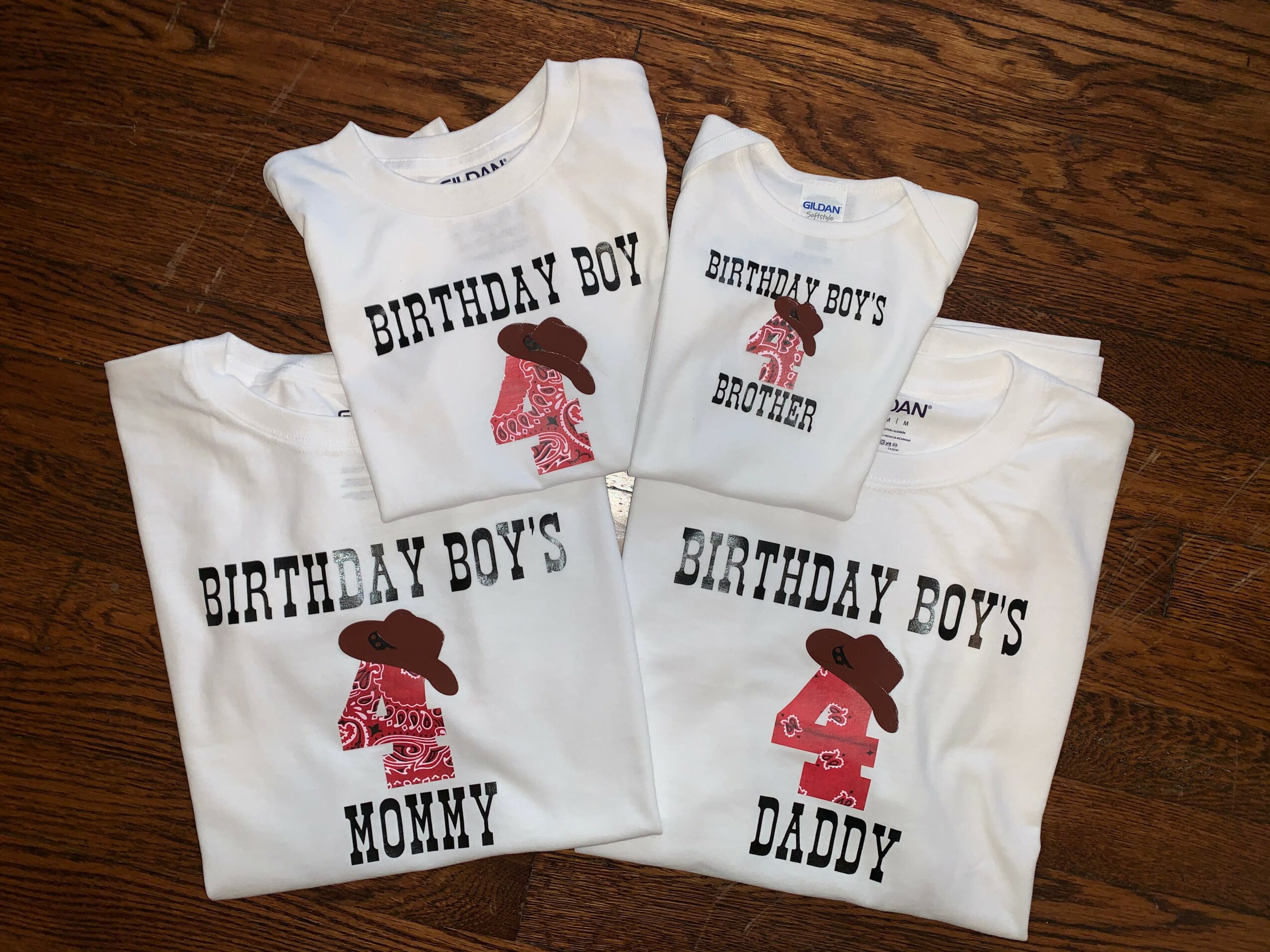 Four white T-shirts with birthday boy themes, featuring a girl with a hat and different titles, on a wooden floor.
