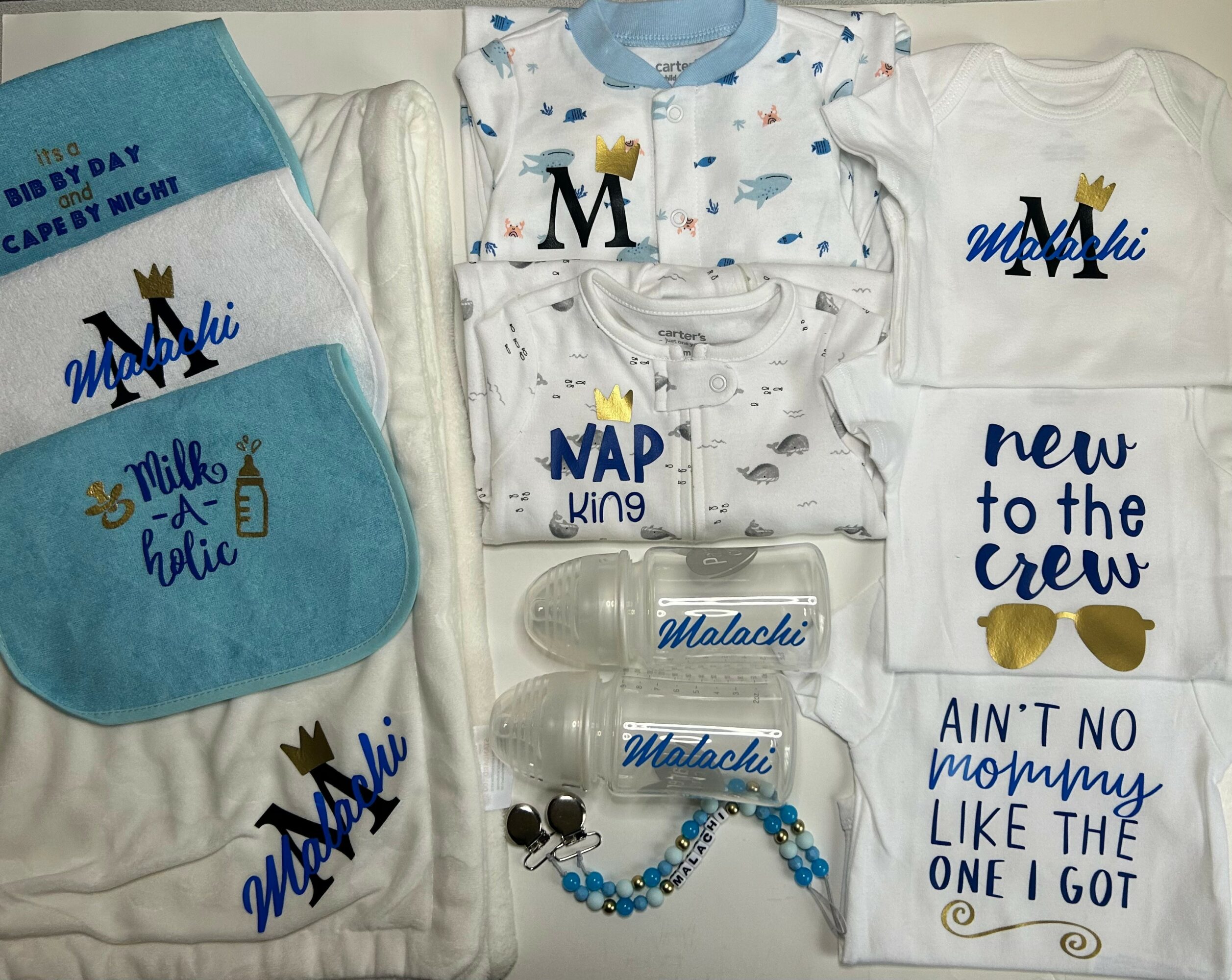 Baby clothing and accessories with blue, white, and gold designs, including towels, onesies, and a bracelet, featuring a crown and text.
