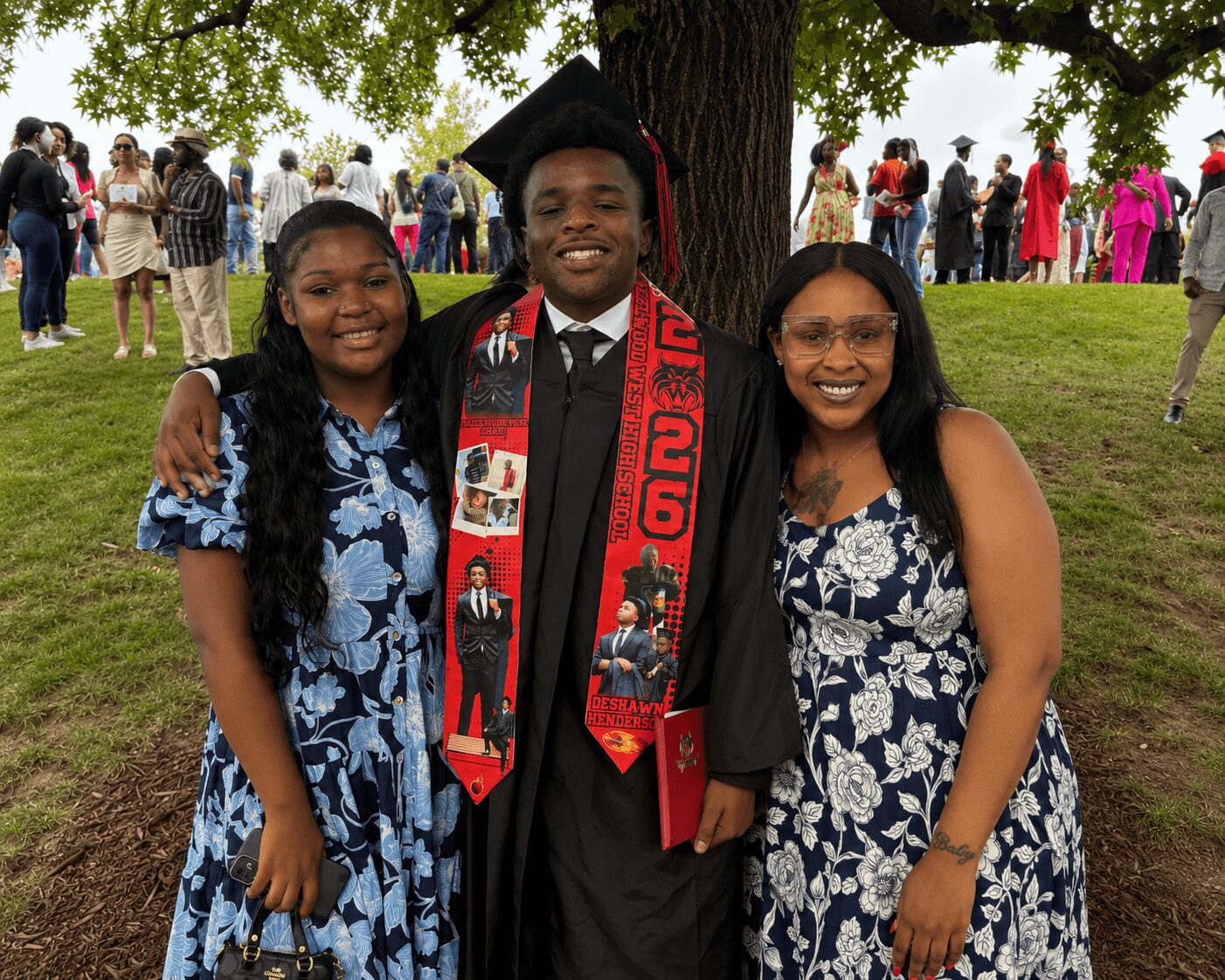 Graduate in cap and gown with two women outdoors, trees and people in background, smiling, holding each other.