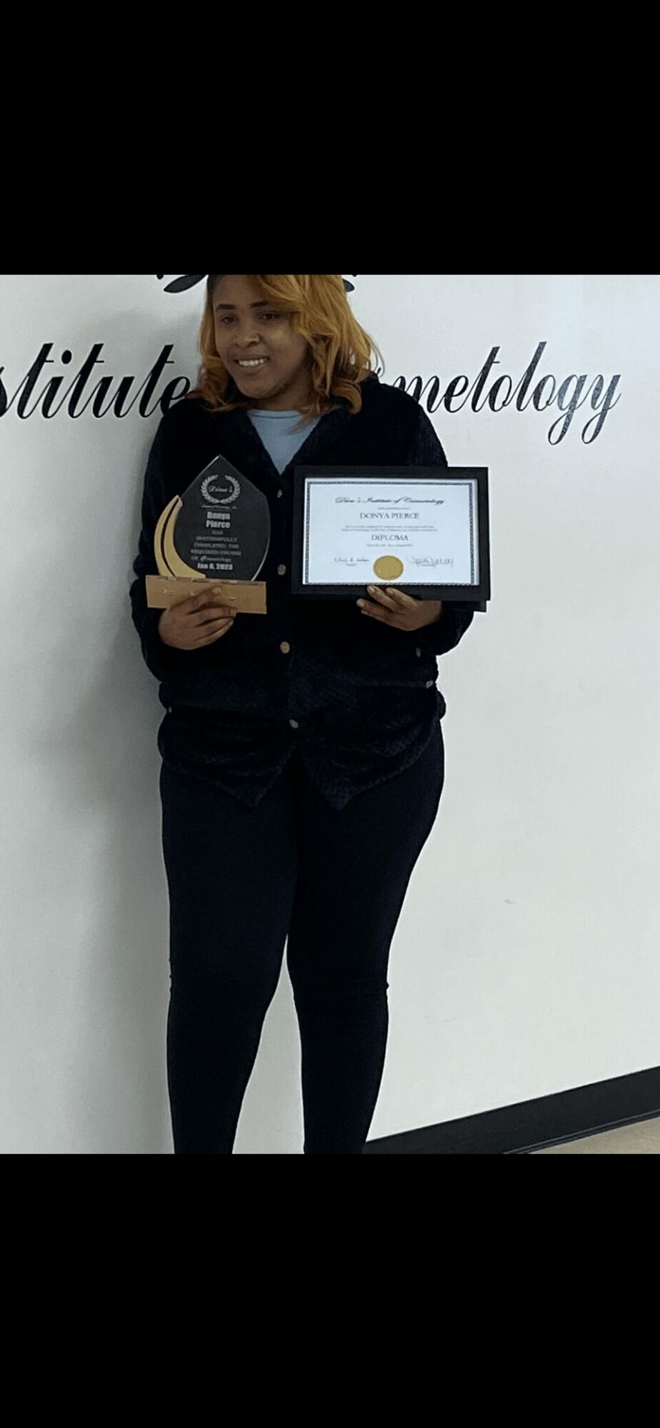 Person holding a trophy and certificate, standing in front of a white wall with text, smiling.