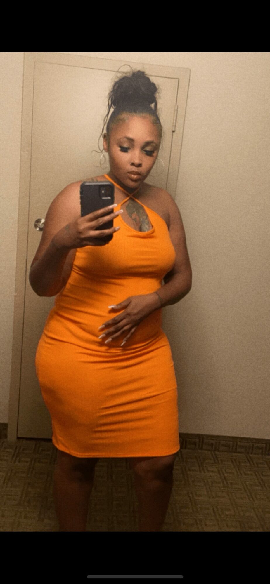 Woman in orange dress taking mirror selfie, standing in front of a door, with her hair in a bun, holding a phone.