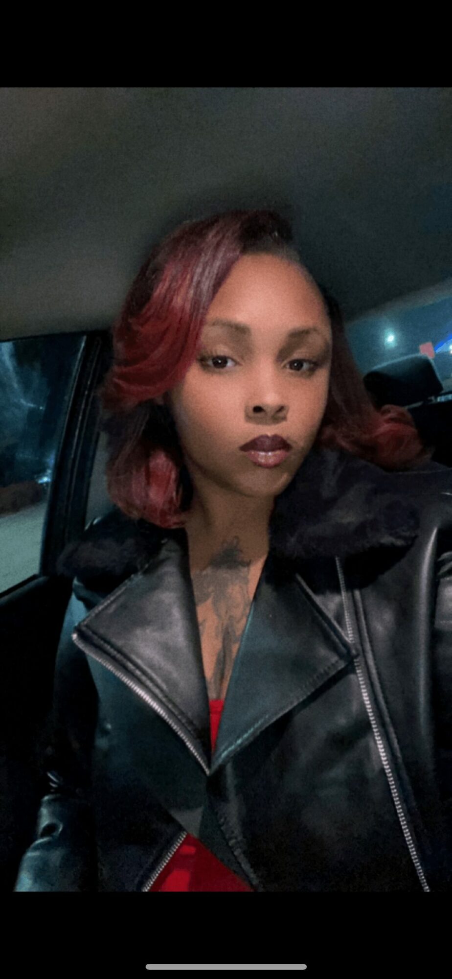 Young woman with red hair in a black leather jacket inside a car, looking at the camera.