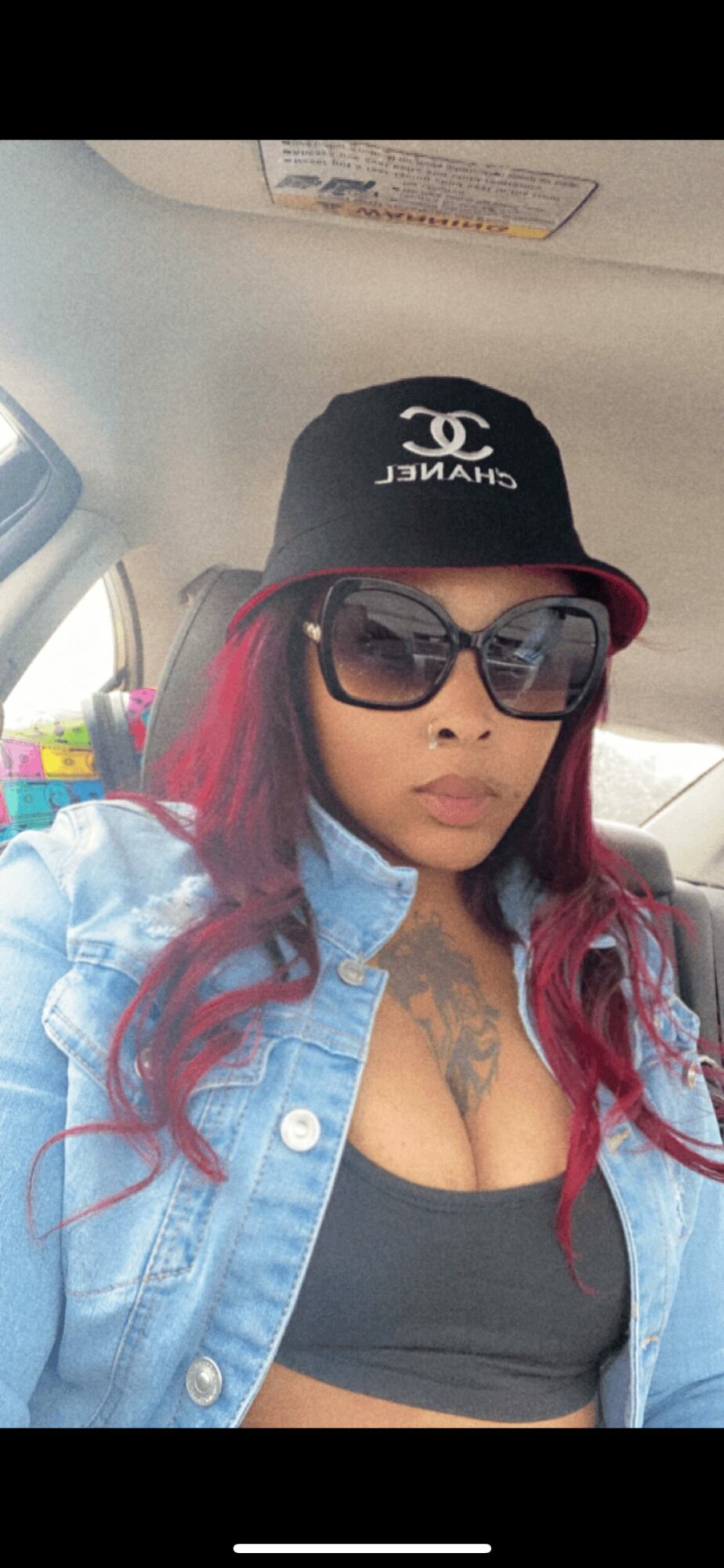 Woman with red hair wearing sunglasses, a black cap, and a denim jacket inside a car.