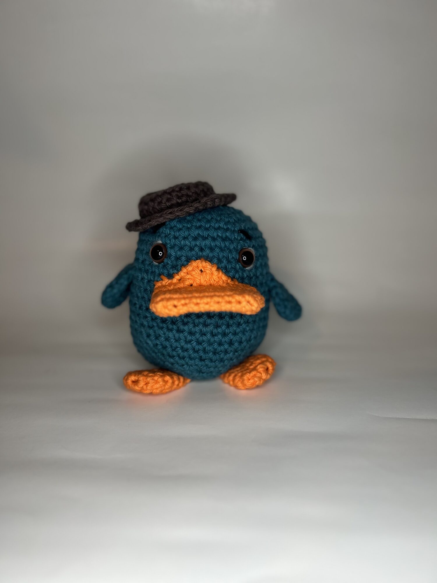Crocheted blue duck with orange beak and feet, wearing a small brown hat, standing on a plain surface.
