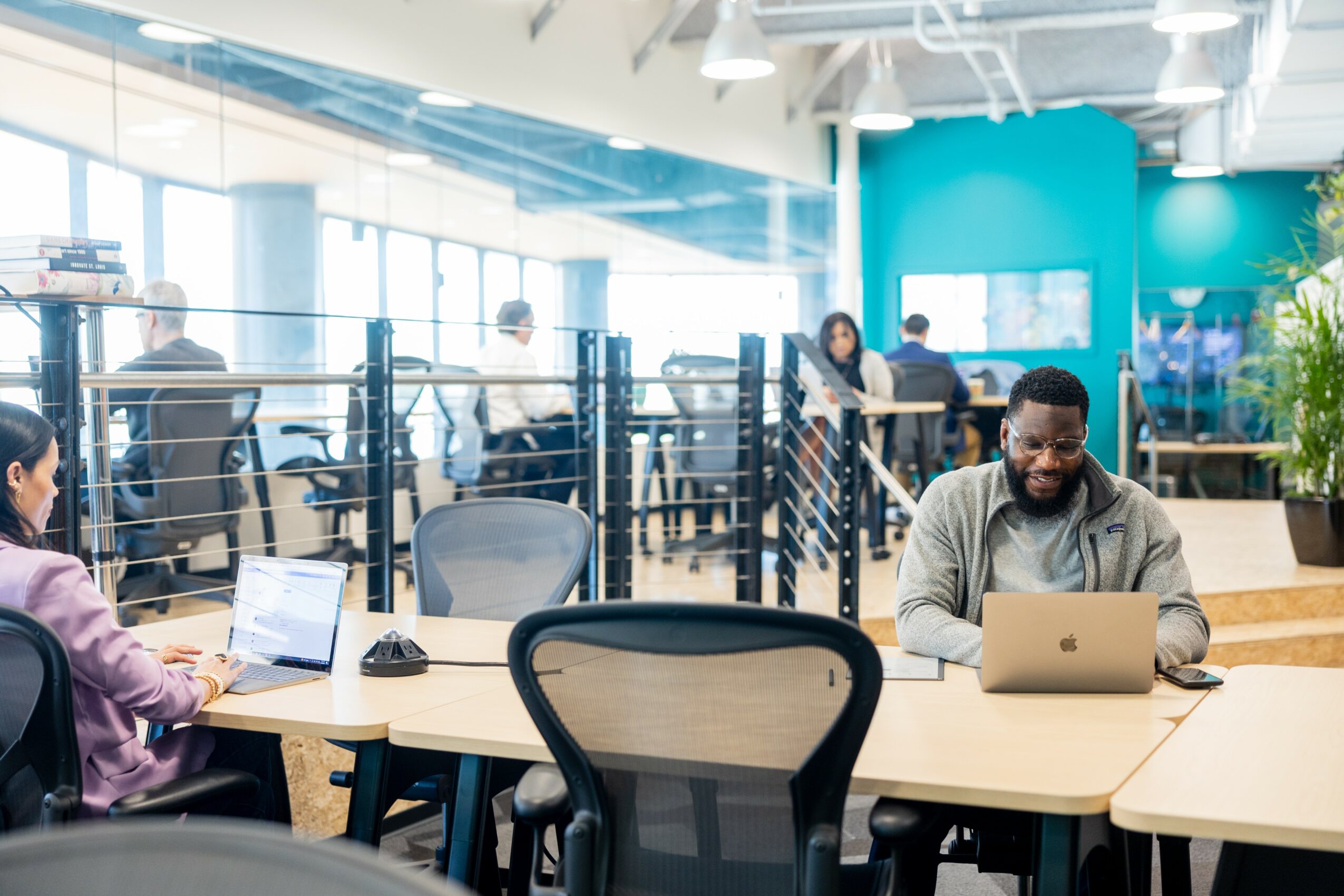 People working in a modern office with large windows, desks, and laptops, some standing and some sitting, with a blue wall in the background.
