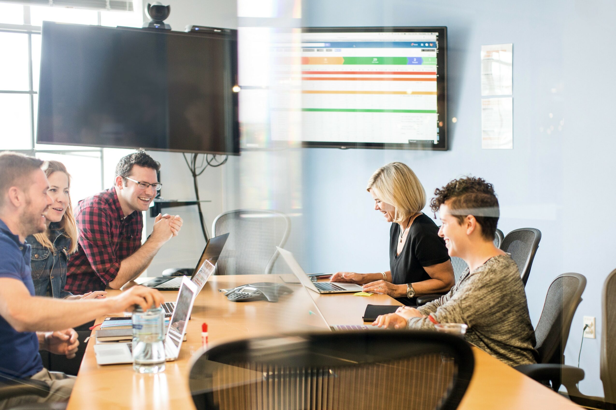 Five people sit around a conference table with laptops, in a modern office with screens on the wall.