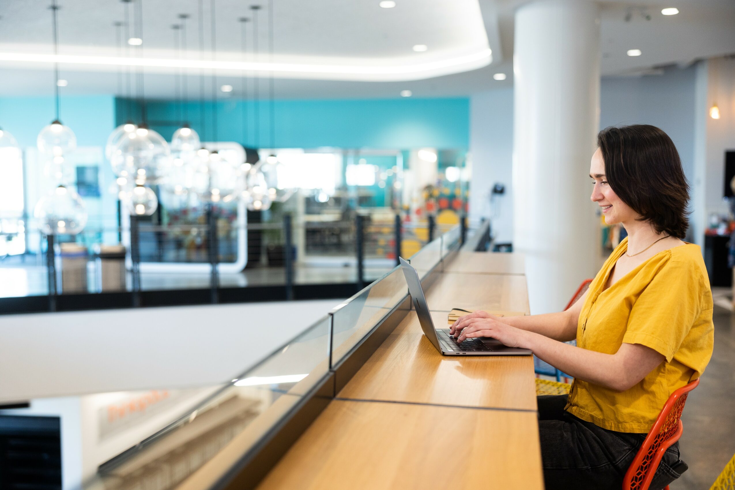 Woman in yellow shirt working on laptop at a wooden counter in a modern indoor space.