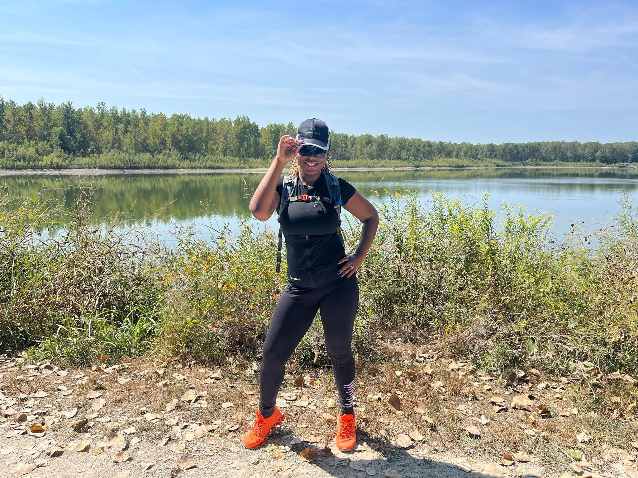 Person standing outdoors near water, smiling, wearing sunglasses, black outfit, orange shoes, and a cap, with greenery and lake in background.