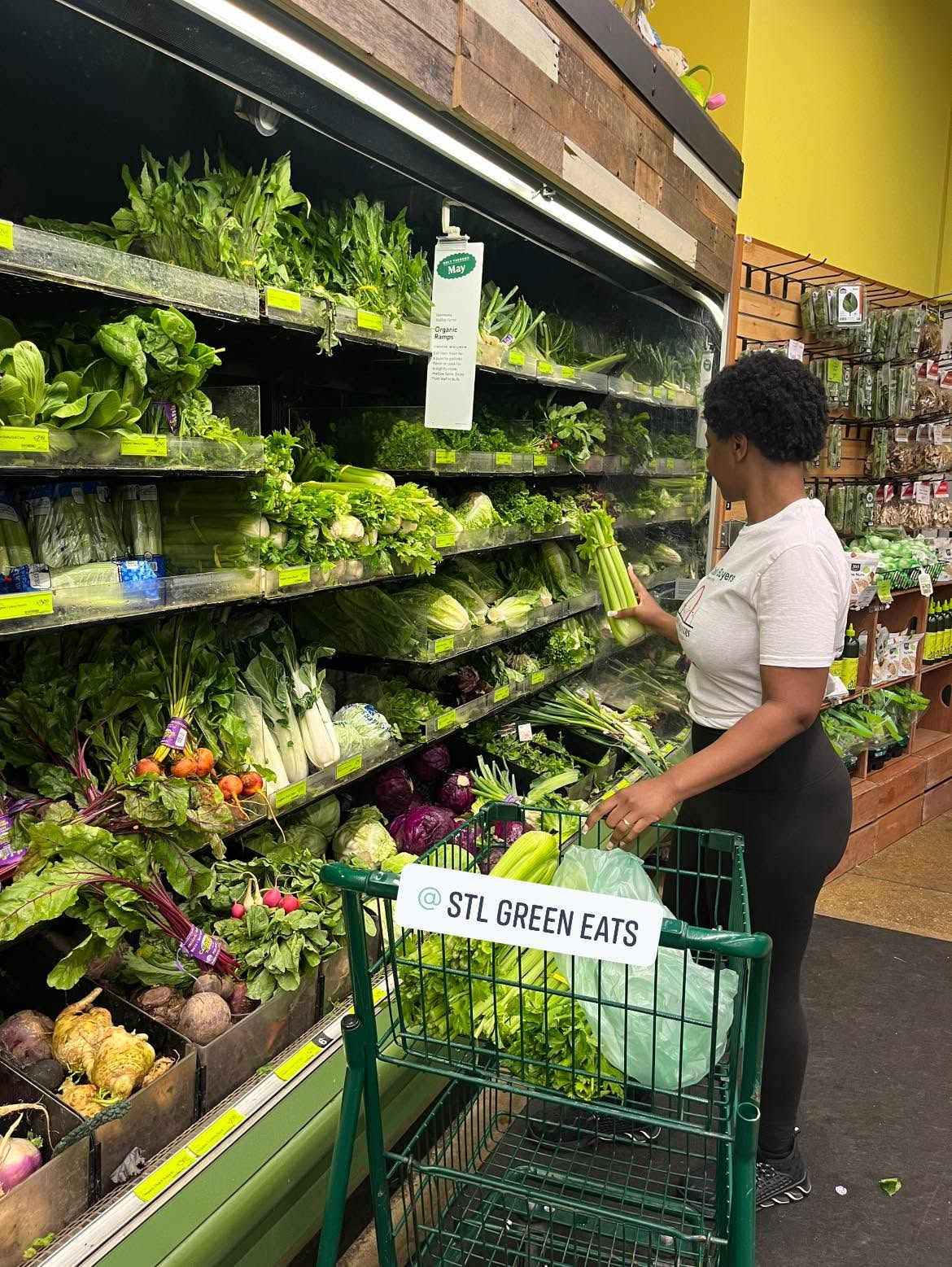 Woman shopping for fresh vegetables in a grocery store, with a cart and shelves of greens and produce behind her.