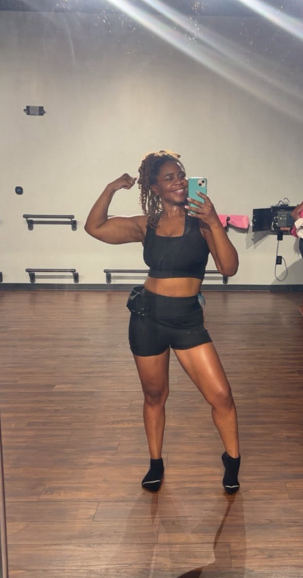 Woman in workout clothes taking a mirror selfie in a dance studio, flexing her arm, with a wooden floor and minimal background.