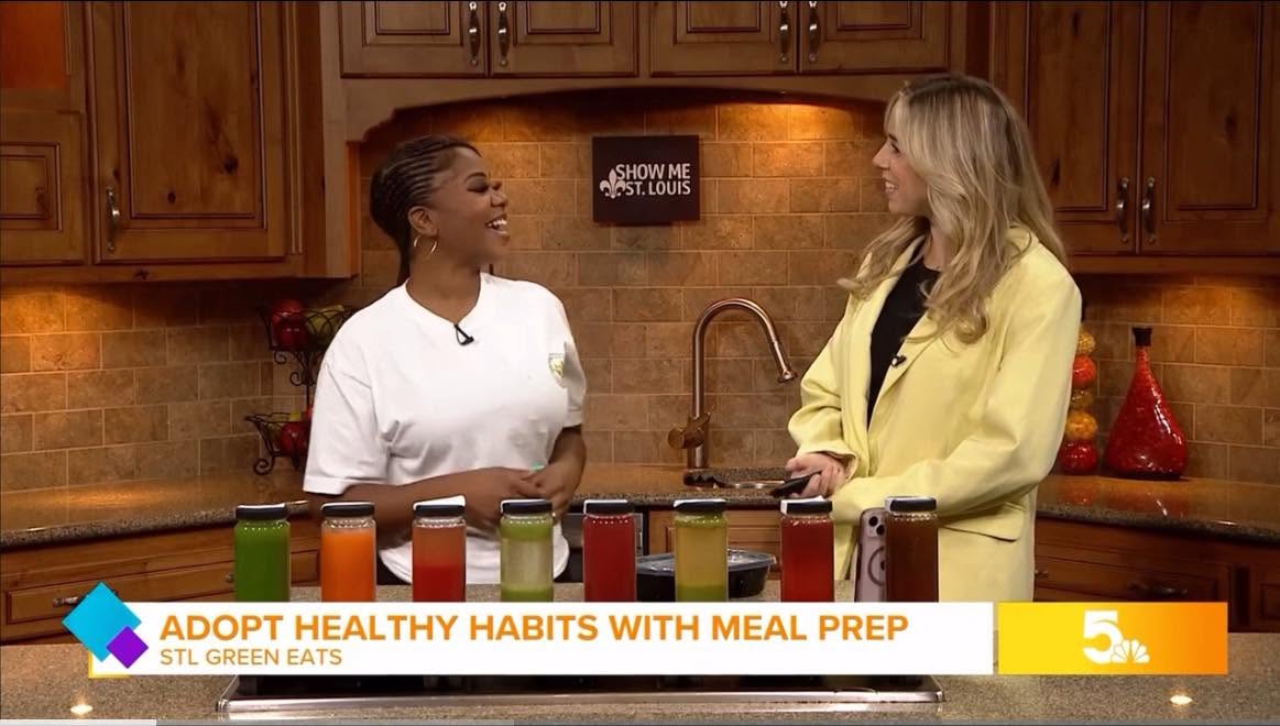 Two women in a kitchen engaged in conversation, with wooden cabinets and a brick backsplash in the background.