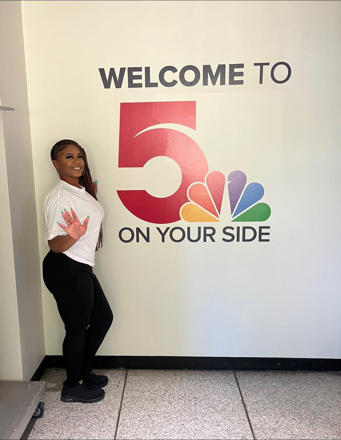 Woman standing next to a wall with a large logo and text, smiling and waving, in a white shirt and black pants.