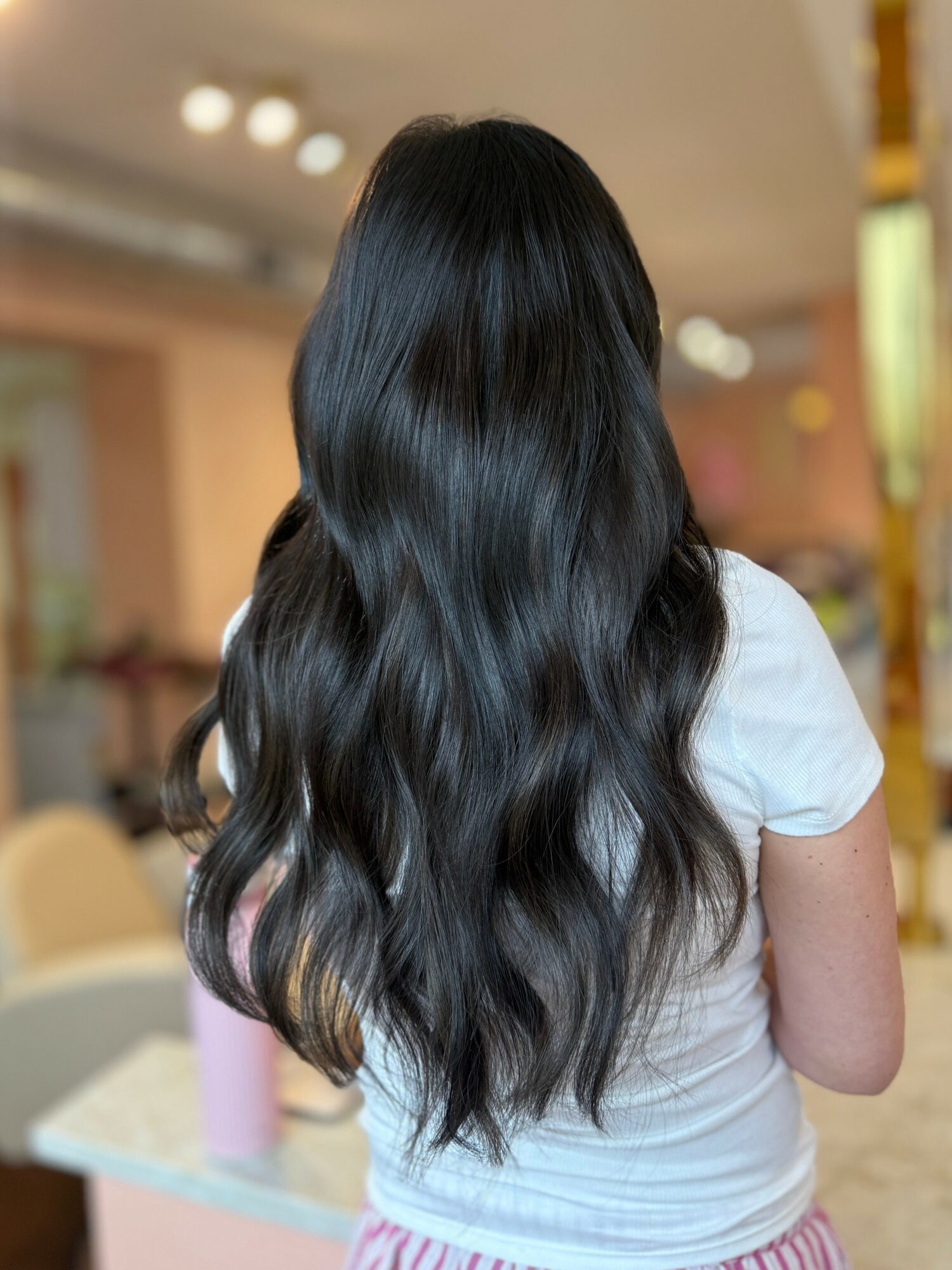 Back view of a person with long, wavy black hair wearing a white shirt in an indoor setting.