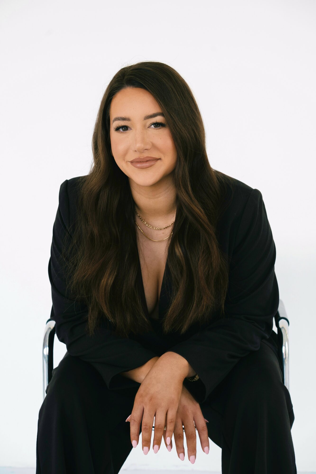 Young woman with long dark hair, wearing a black blazer, sitting with hands on her knees, smiling softly, against a white background.