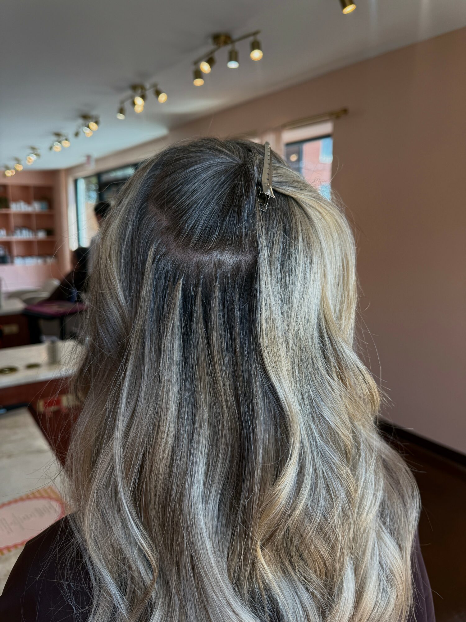 Back of a person's head with long, wavy, blonde hair in a room with windows and ceiling lights.