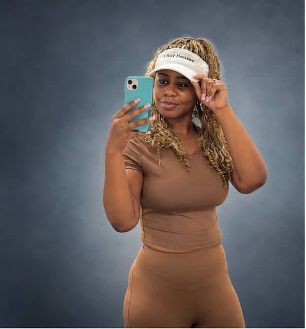 Woman with curly hair wearing a visor and brown outfit, taking a selfie against a dark background.
