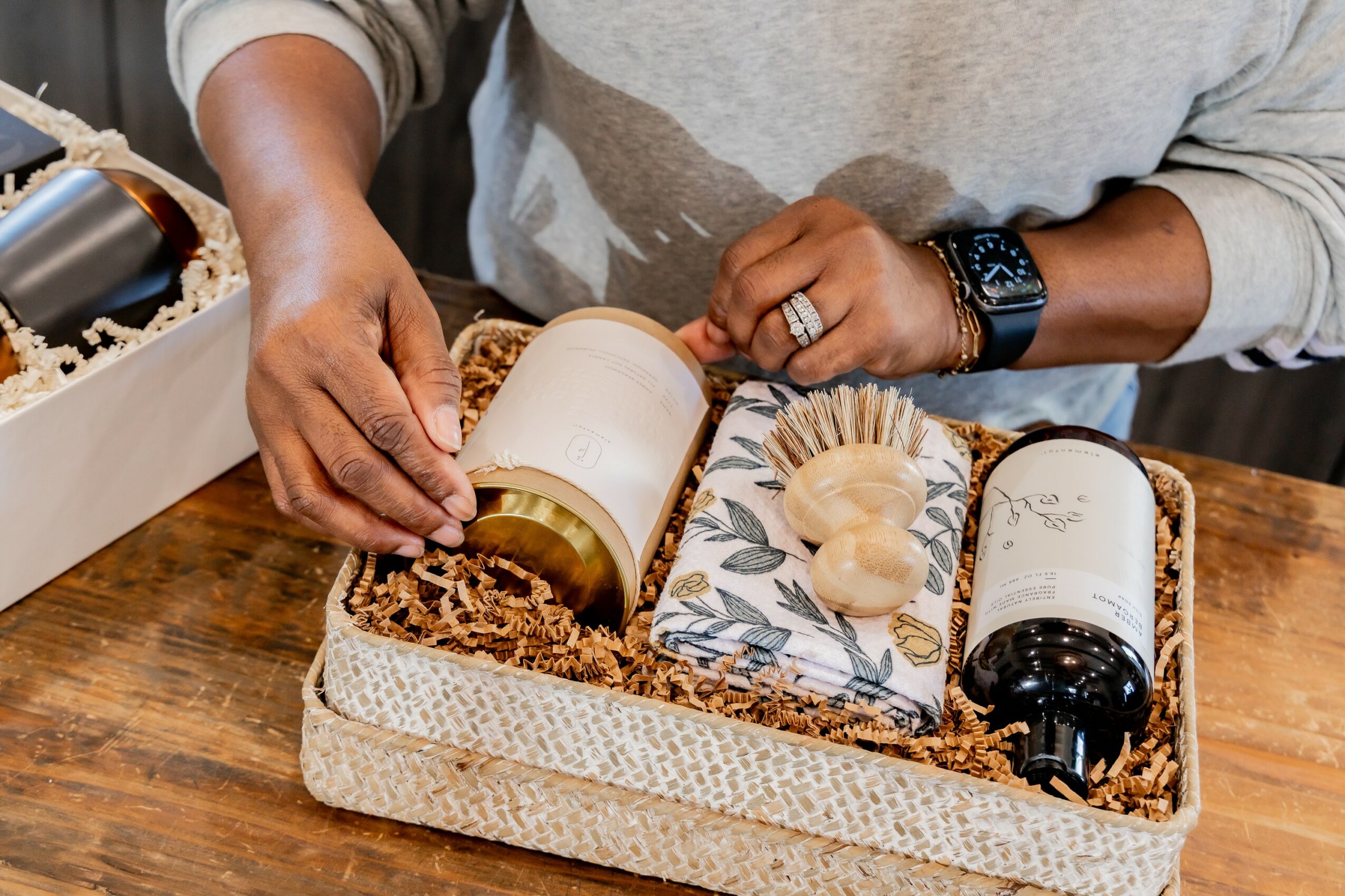 Person opening a gift box with candles and decorative items inside, on a wooden surface.