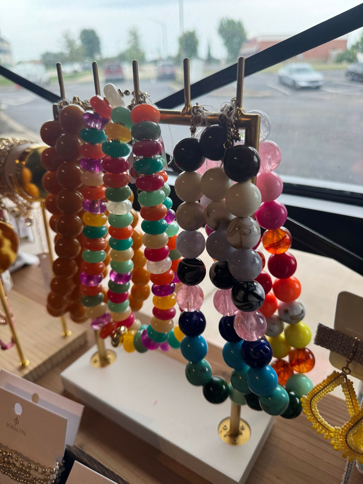 Display of colorful beaded jewelry on a white surface near a window, with a blurred outdoor scene in the background.