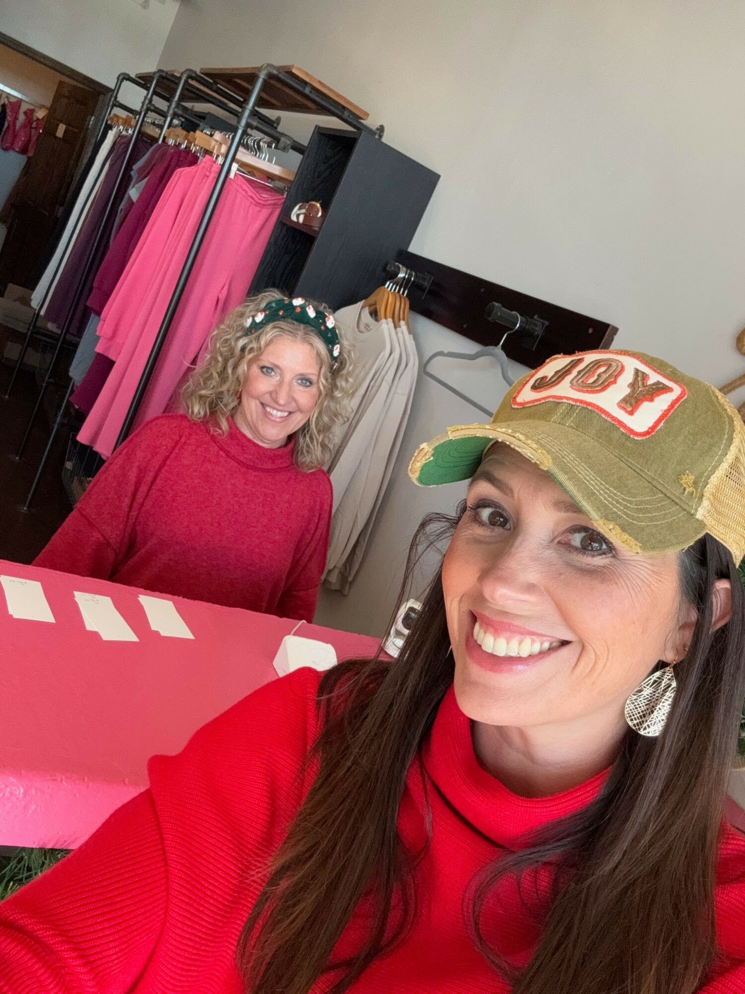 Two women smiling, one in a red sweater and cap, the other in a pink sweater with a headband, in a room with clothing racks.