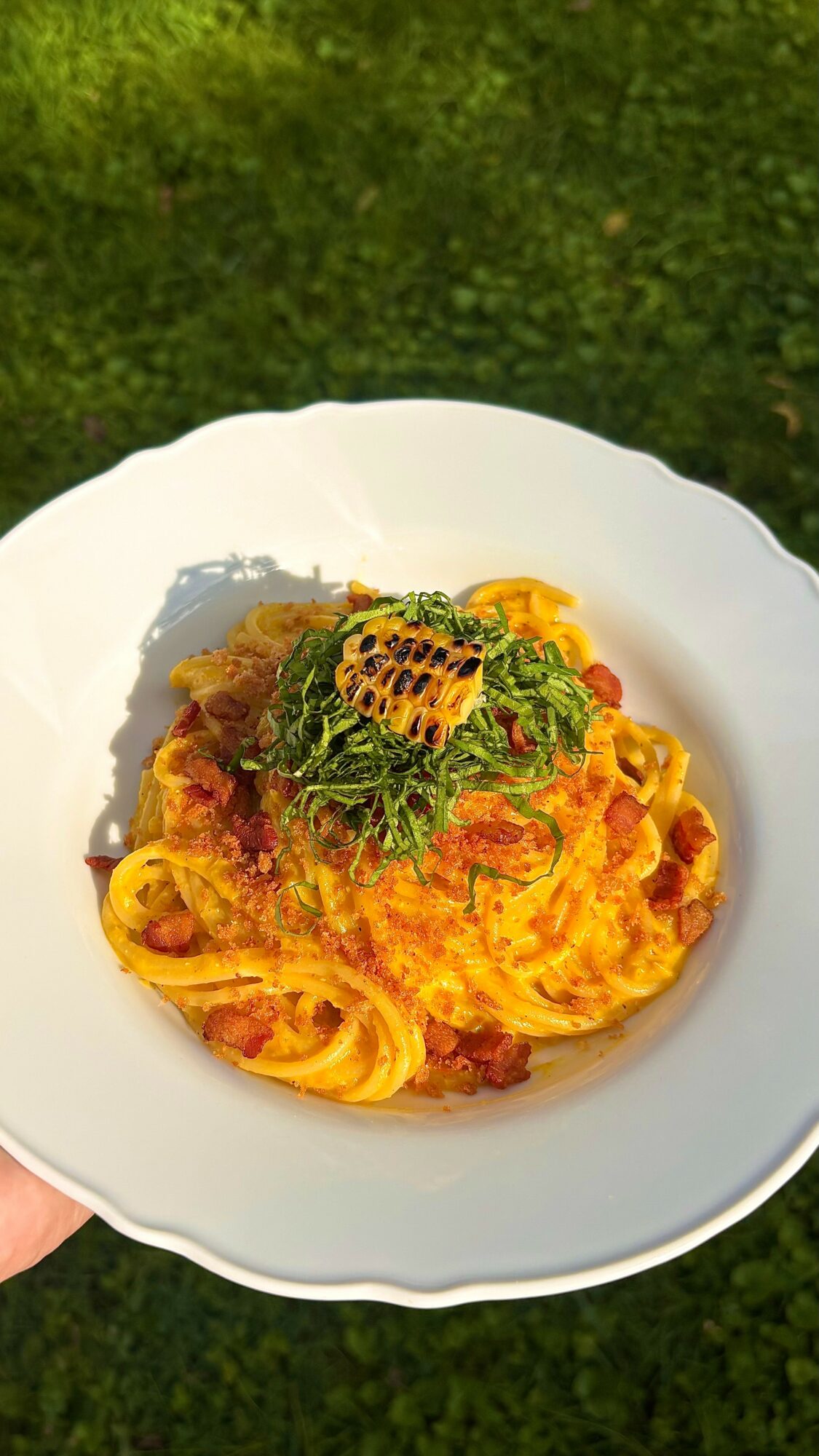 Plate of pasta with sauce, topped with green herbs and a yellow flower, held outdoors against a grassy background.