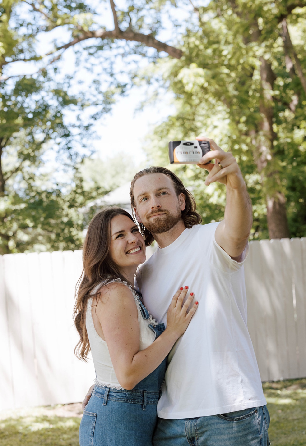 A couple takes a selfie outdoors with trees and a white fence in the background.