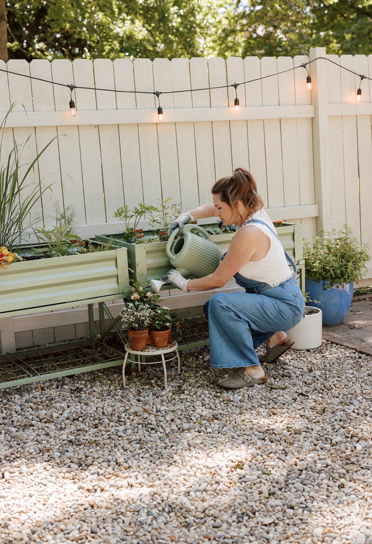 Woman watering plants in a backyard garden with string lights, a white fence, and potted plants, sitting on a white stool.