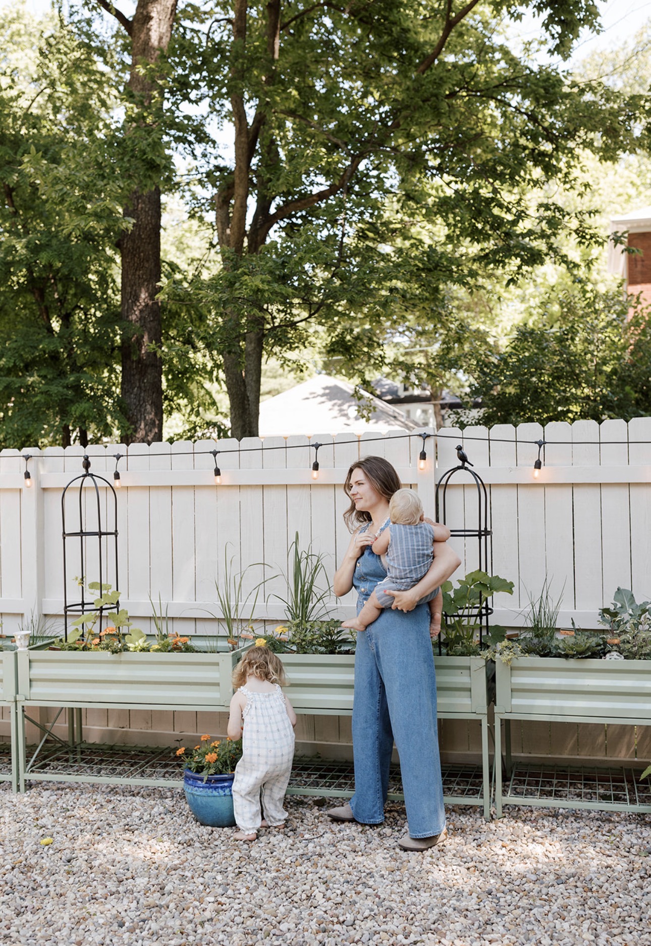 Woman holding a child and a toddler with a blue bucket in a backyard with trees and a white fence.