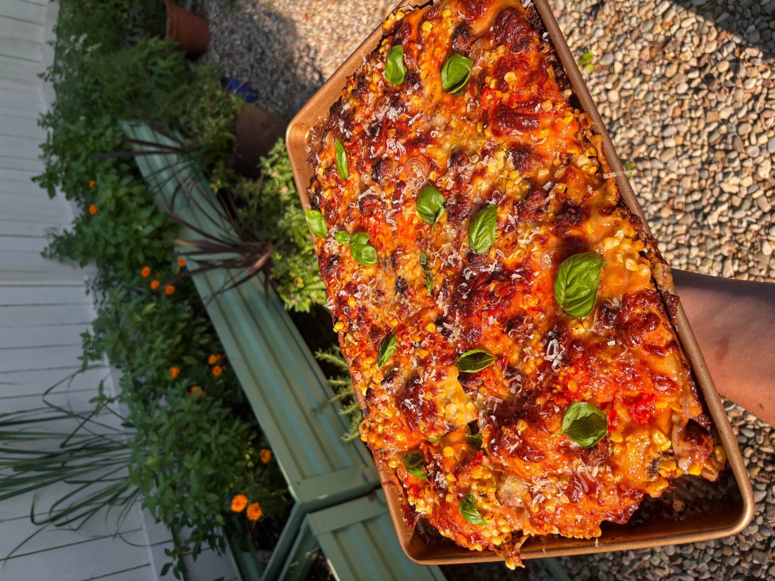 A rectangular pizza with cheese, tomato sauce, and basil leaves on top, placed on a tray outdoors.