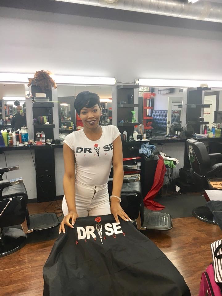 Woman with short hair and white dress standing behind a table in a hair salon, smiling, with salon mirrors and chairs in background.