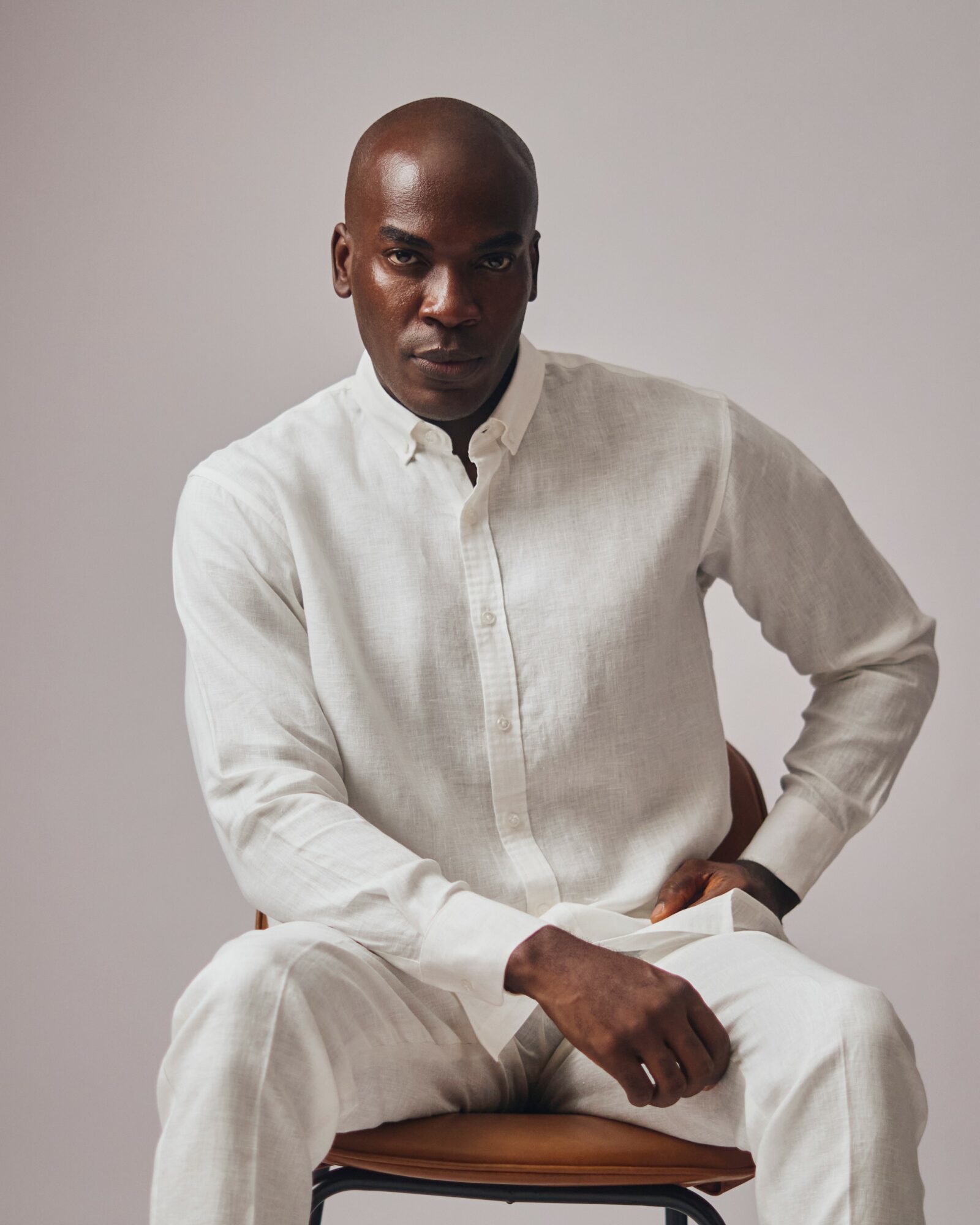 A person with a shaved head wearing a white long-sleeve shirt and matching pants, sitting on a chair against a plain background.