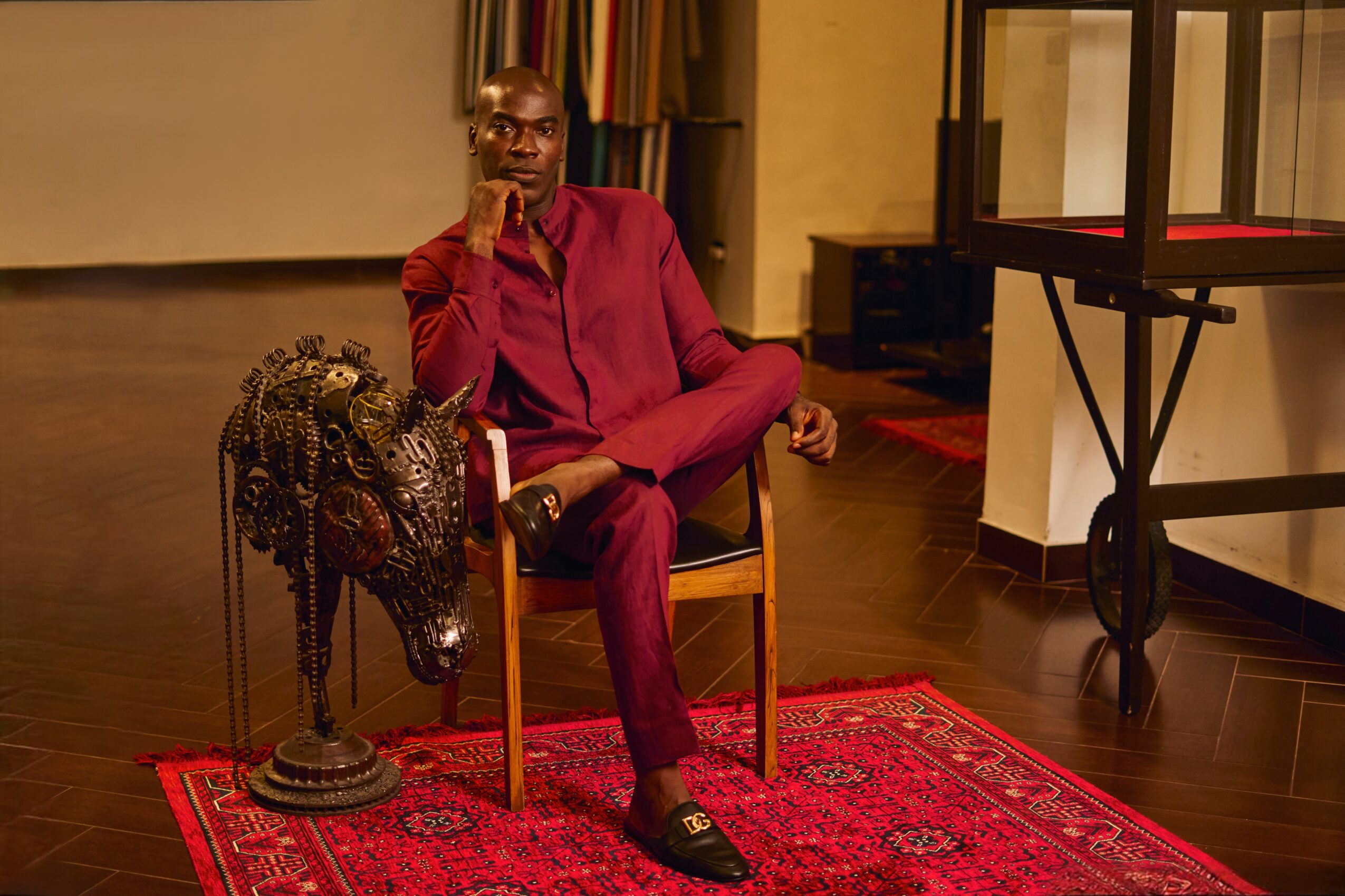 Man in a maroon suit sitting on a wooden chair on a red patterned rug, with a decorative object nearby.