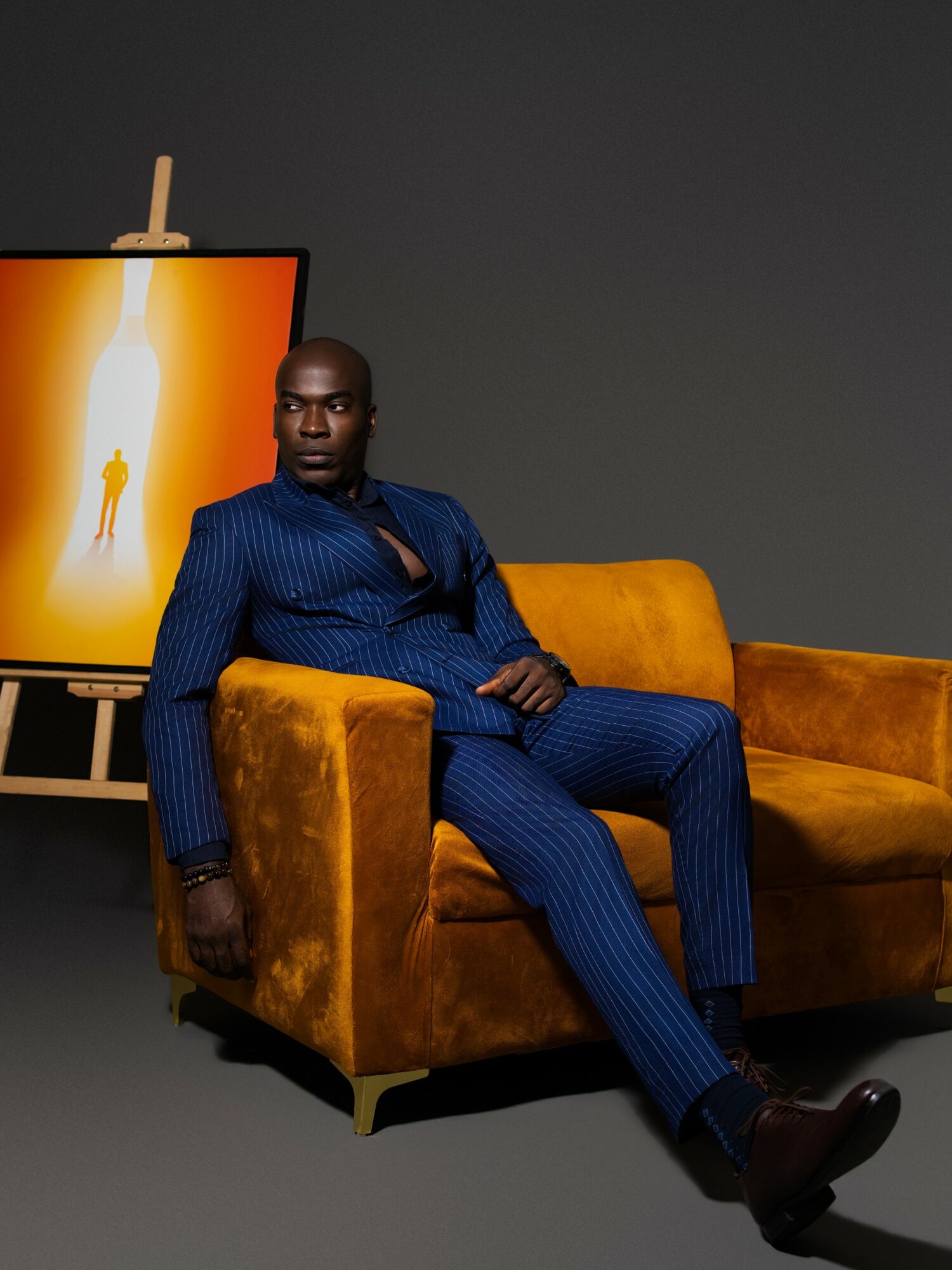 Man in a blue pinstripe suit sitting on a yellow velvet sofa, with a painting of a silhouette in the background.
