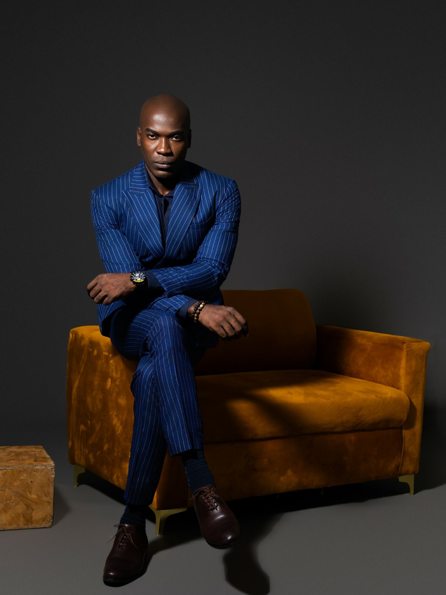 Man in a blue pinstripe suit sitting on a mustard-colored sofa with a dark background.