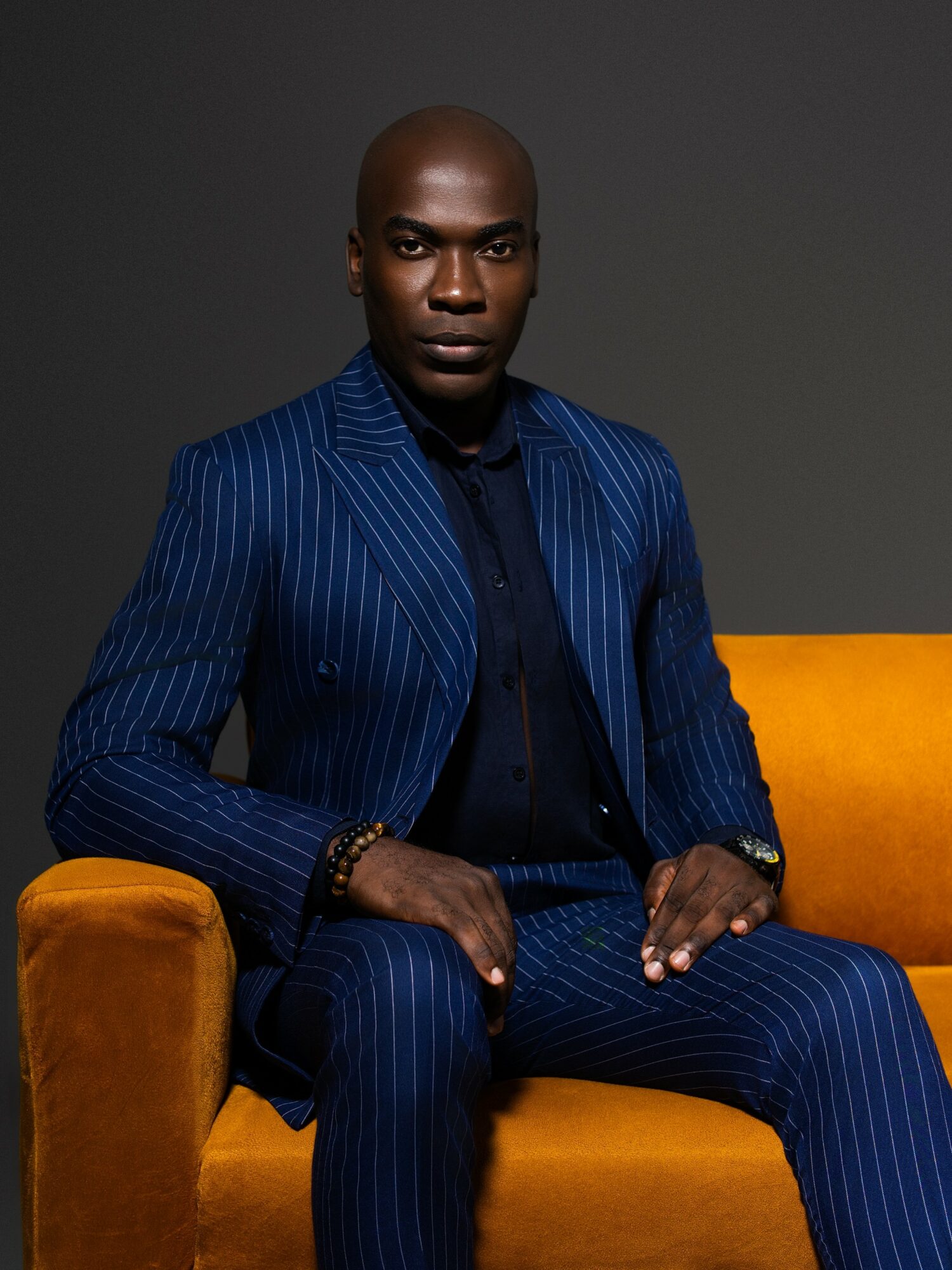 Person with a shaved head wearing a blue pinstripe suit, sitting on a yellow couch against a dark background.